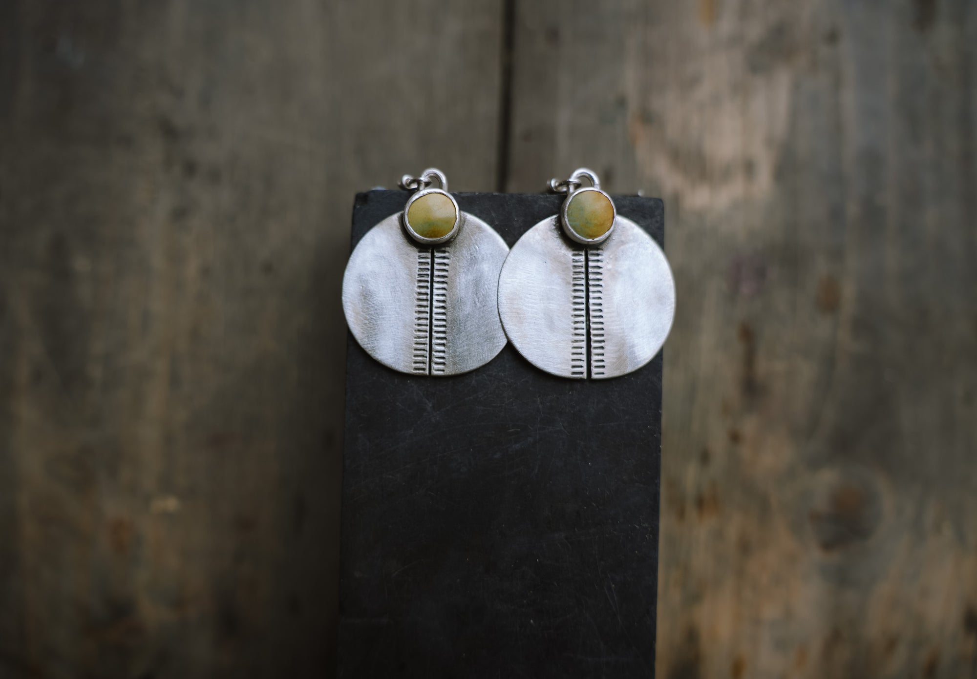 A pair of hand-stamped sterling silver circular earrings with green turquoise gemstones on a dark background.