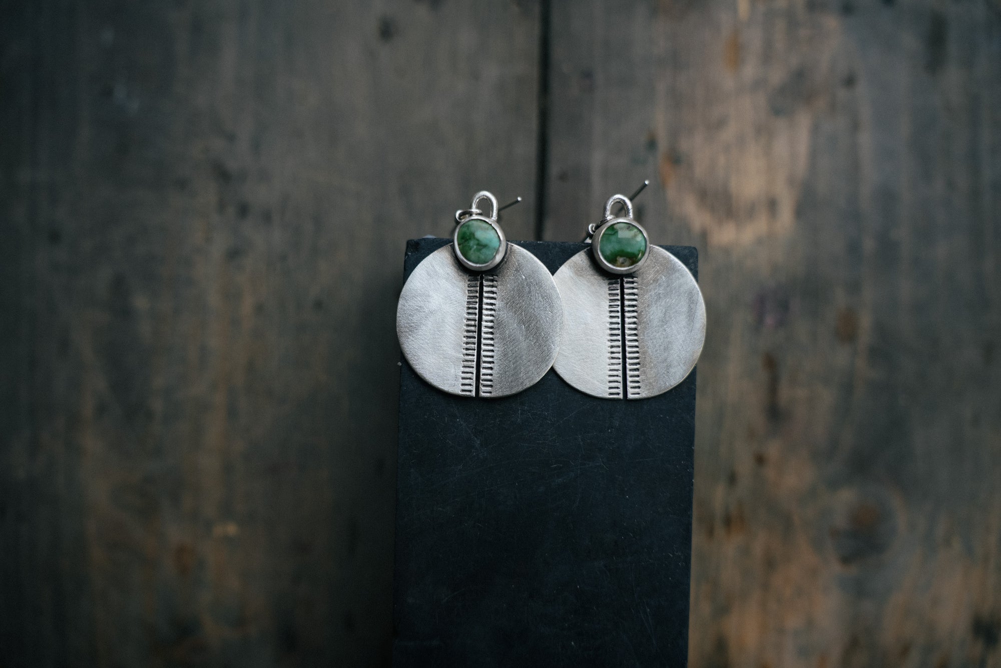 A pair of hand-stamped sterling silver circular earrings with green/blue turquoise gemstones on a dark background.