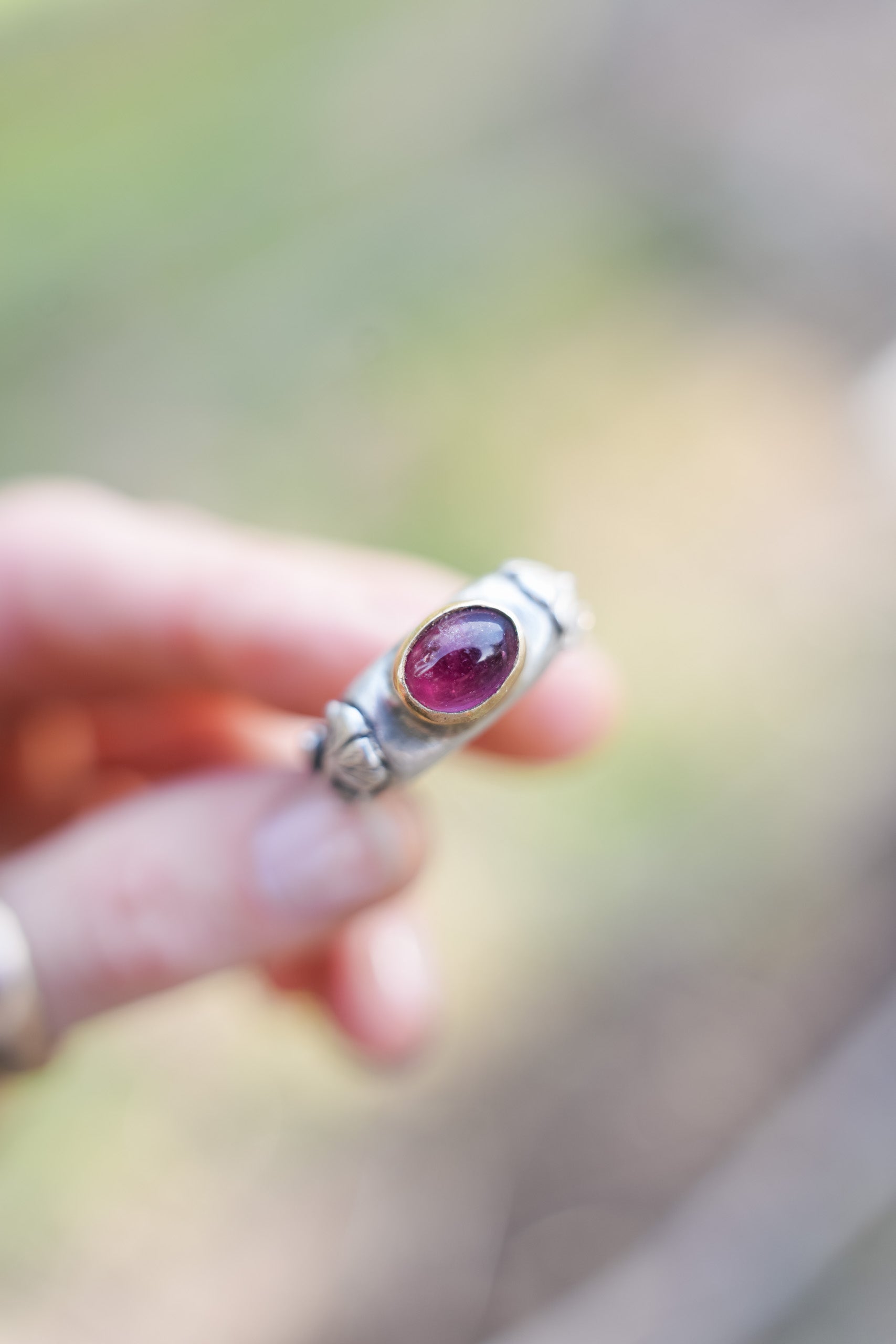 A woman's hand holding a sterling silver ring with a pink tourmaline gemstone. The gemstone is set in an 18K gold bezel, with hand-carved silver poppy flowers on the sides of the band. The natural background is blurred. 