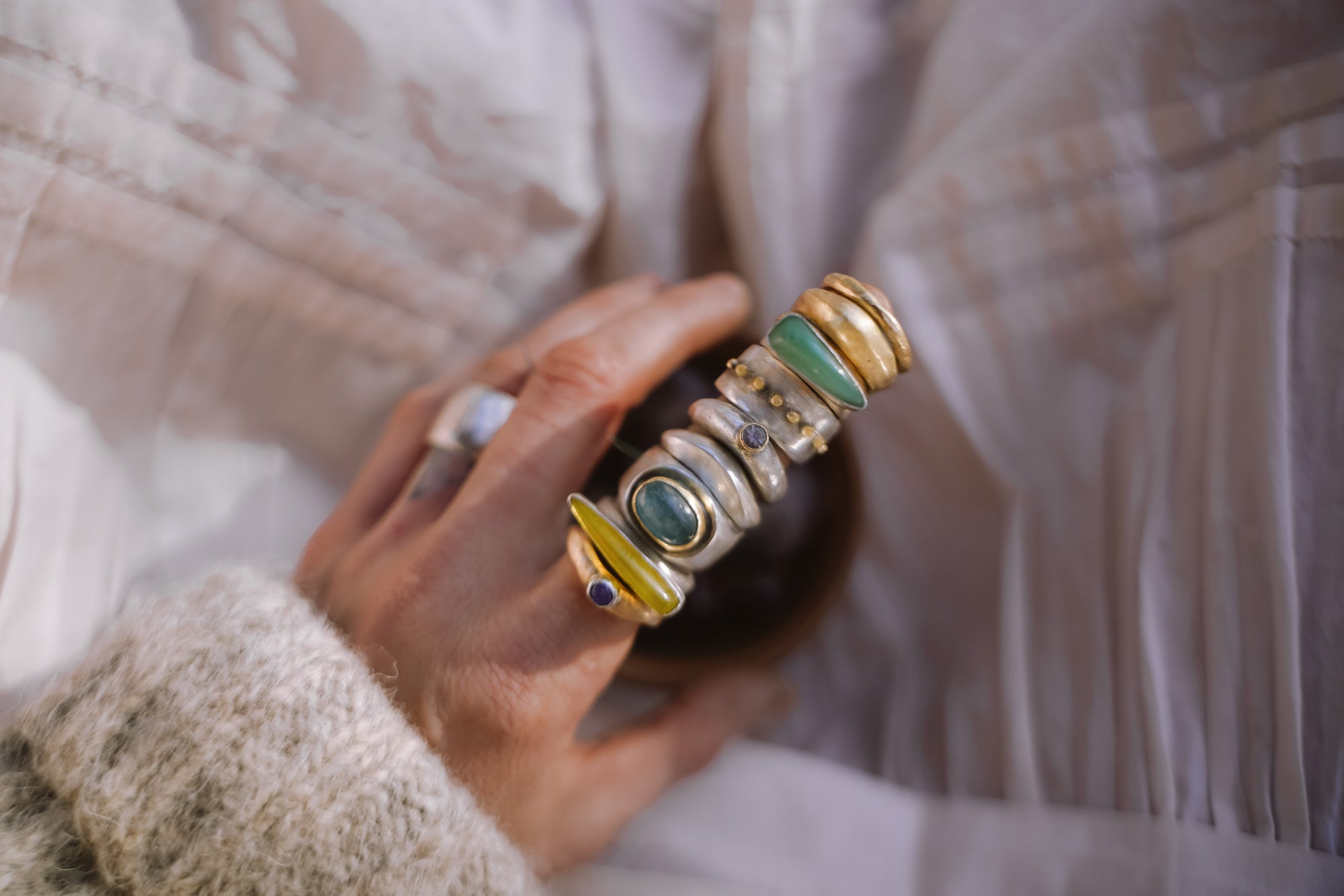 A woman's hand against a fabric background. She is wearing a stack of rings on her forefinger. They are different designs: some are sterling silver, some are silver and gold, and some include gemstones.