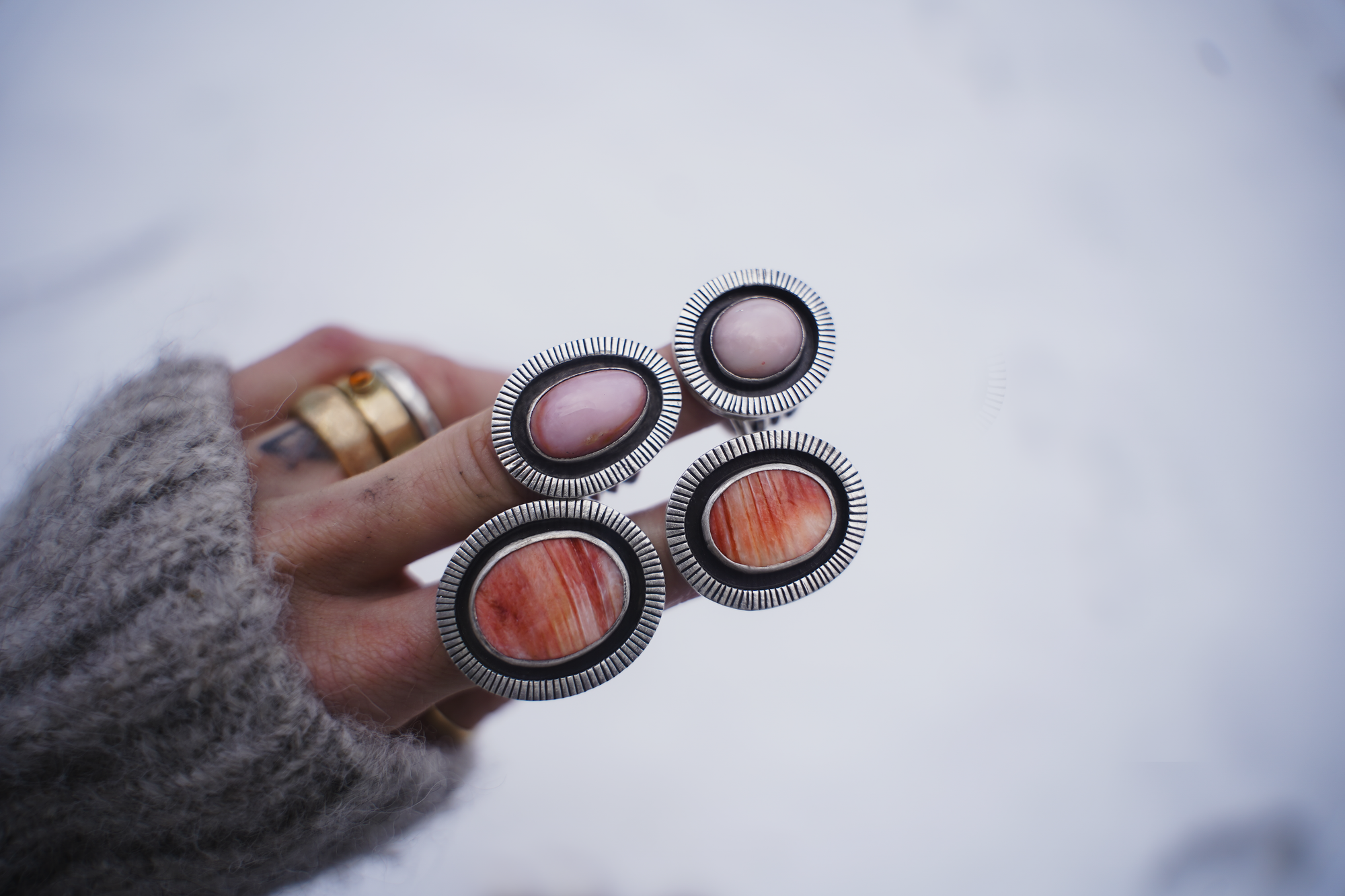 A woman's hand with stacks of bold and chunky sterling silver and gemstone rings on two of her fingers. The natural background is blurred.