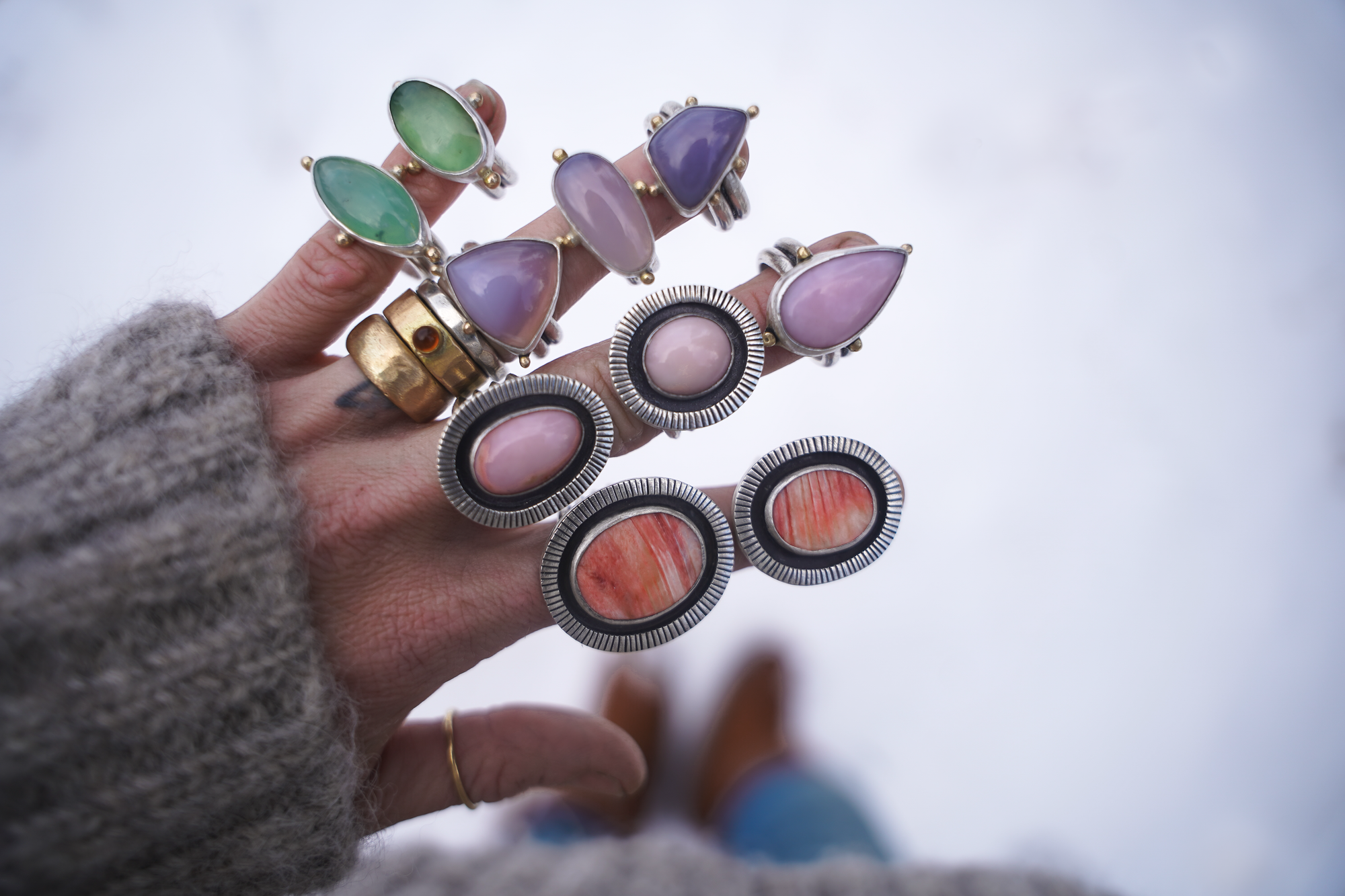 A woman's hand with stacks of bold and chunky sterling silver and gemstone rings on each of her fingers. The natural background is blurred.