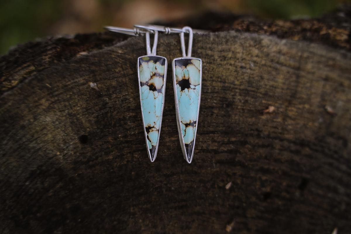 A pair of sterling silver dangler earrings with triangular turquoise pendants, resting on a dark, textured wood surface.