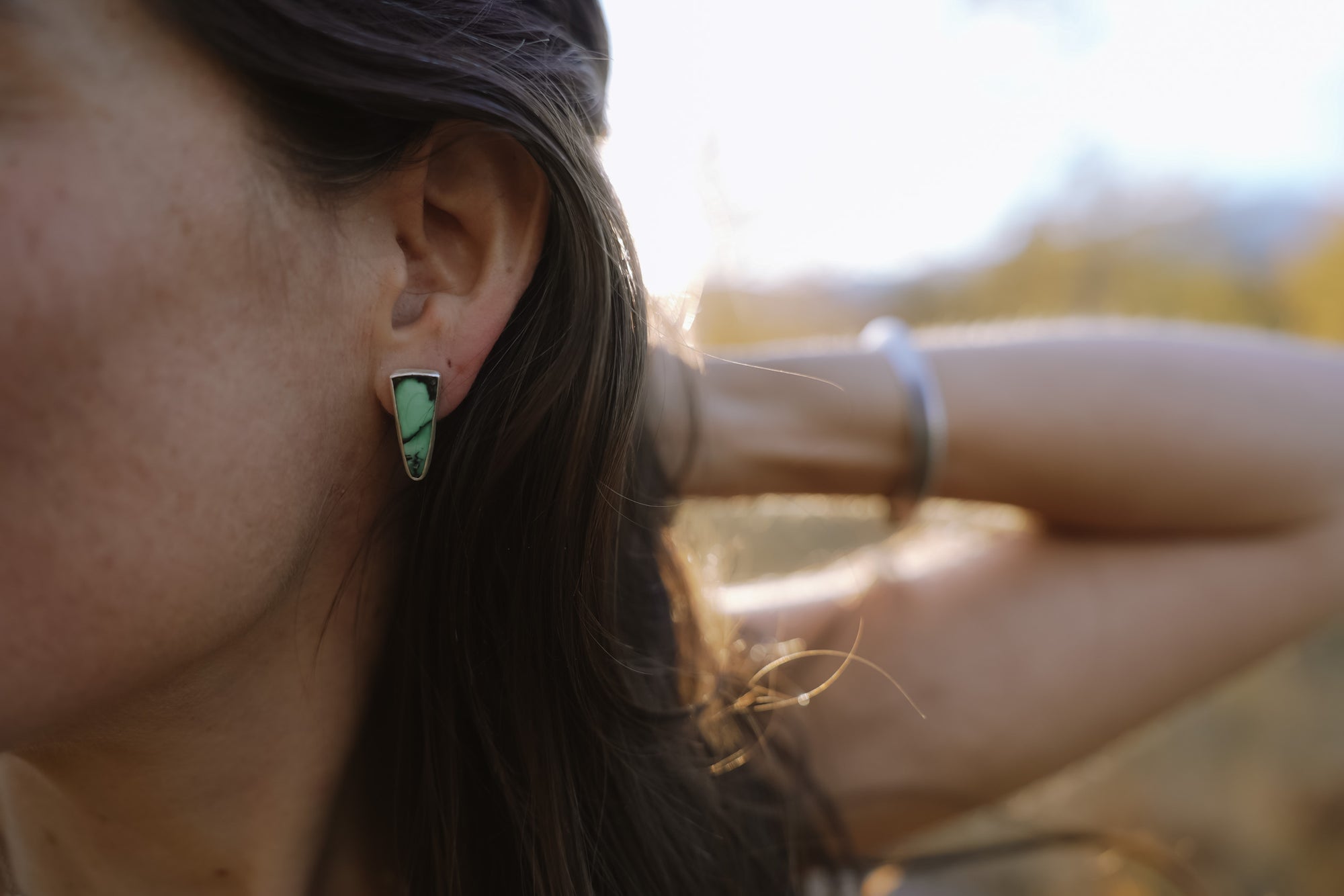 Close-up of a woman wearing a silver and green gemstone spiked-shaped post earring with a blurred natural background.