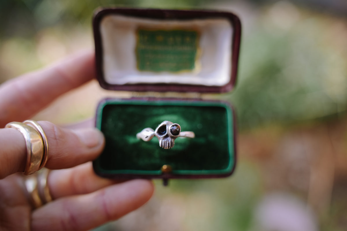 A sterling silver skull ring set with black garnet, displayed in an open vintage green velvet ring box held by a woman's hand.