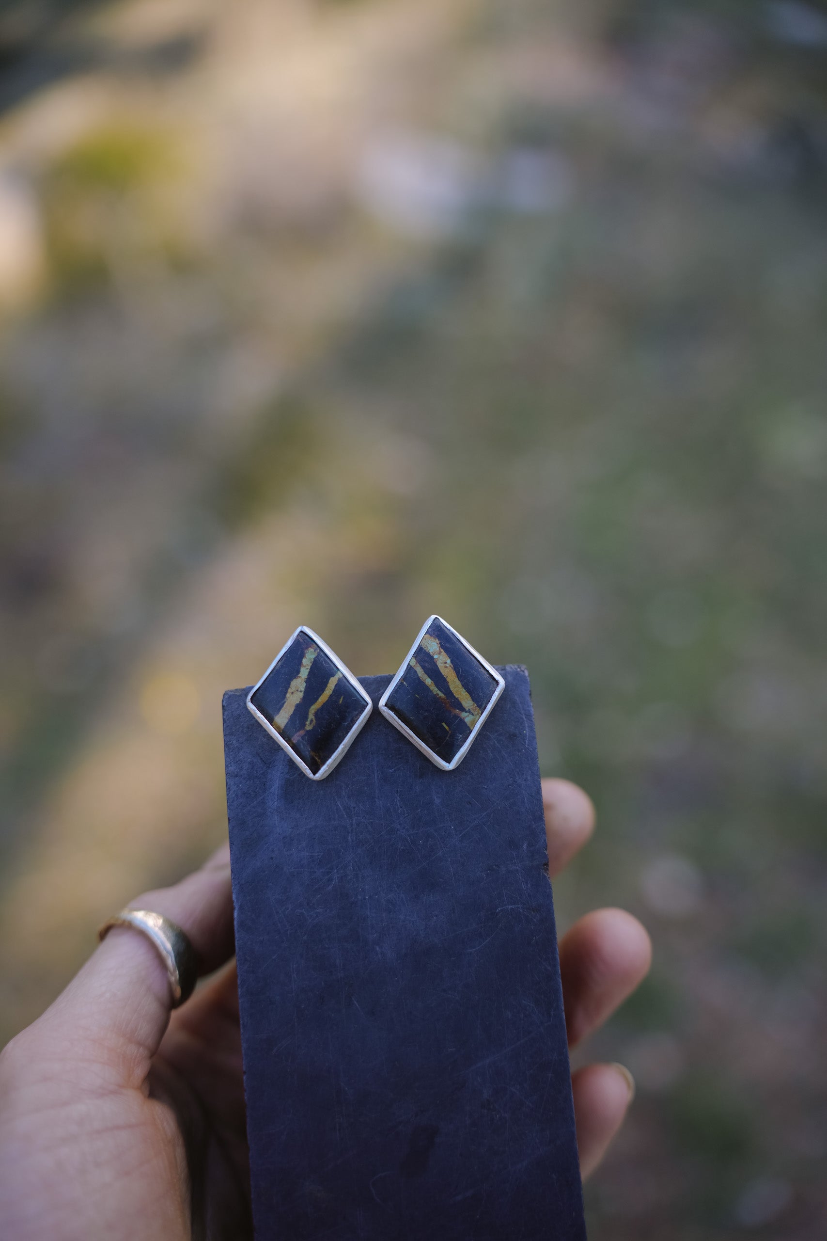 A woman's hand holding a blue velvet rectangle onto which a pair of sterling silver and blackjack post earrings are attached. The earrings are diamond shaped.