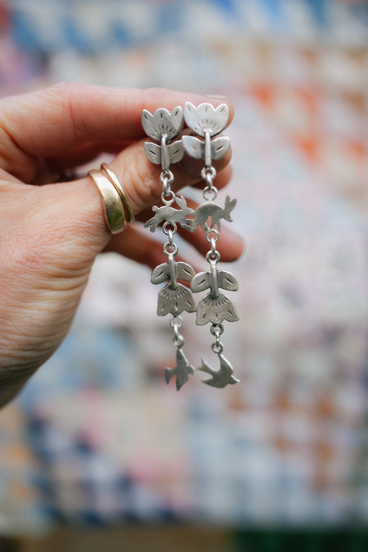 A woman's hand holding a pair of silver dangle earrings with birds, flowers, and rabbits, set against a blurred multicolored background.
