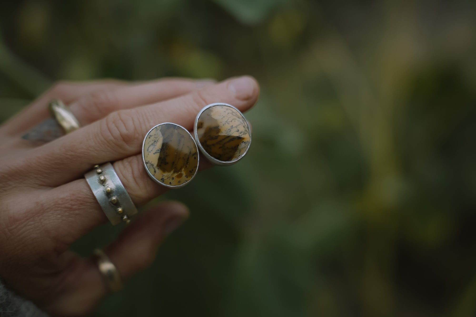A woman's hand holding a pair of sterling silver post earrings with a round rich mustard-yellow and brown-orange gemstones against a blurred natural background.