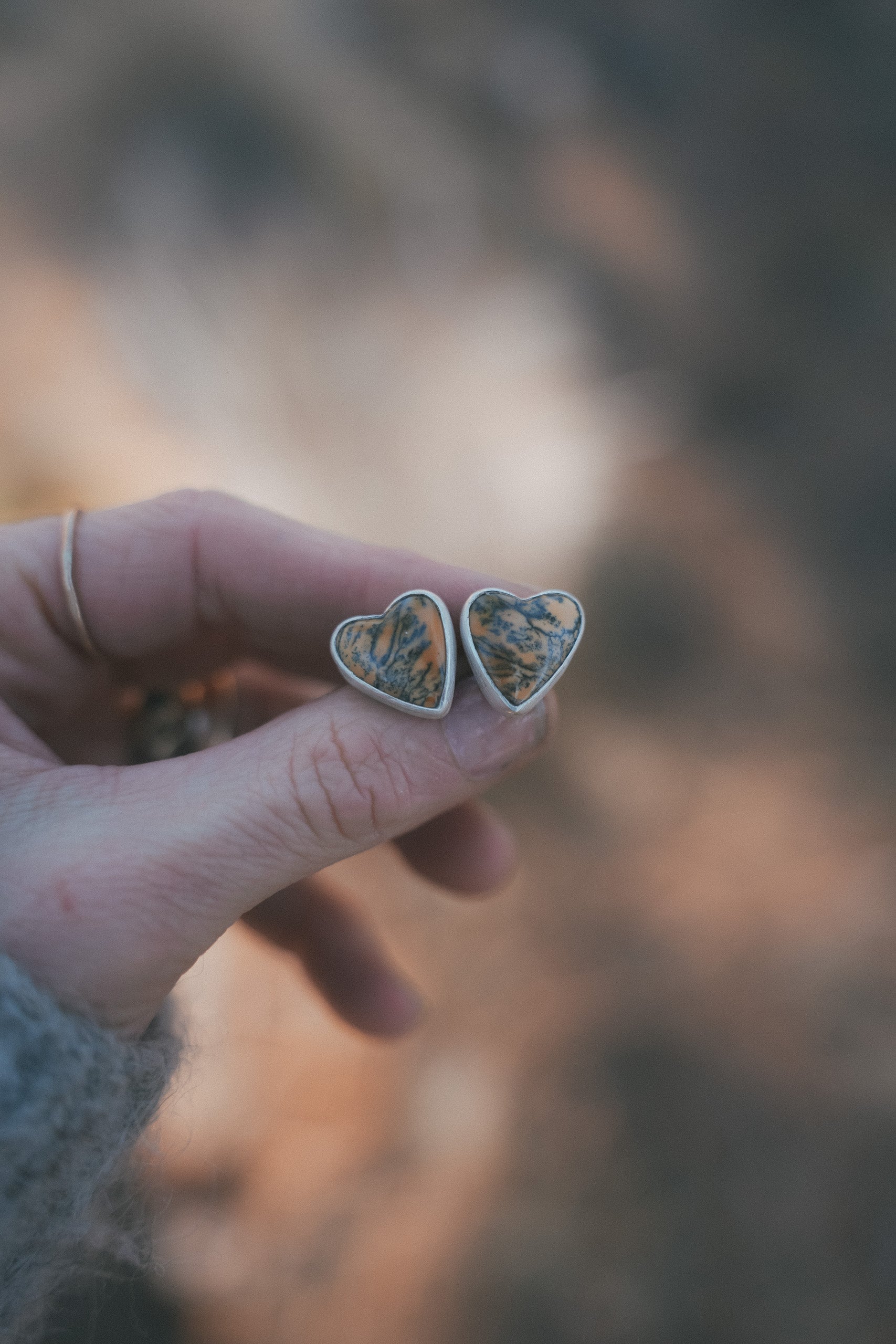 Heart-shaped sterling silver and yellow-orange dendritic jasper earrings held in a woman's hand against a blurred natural background.