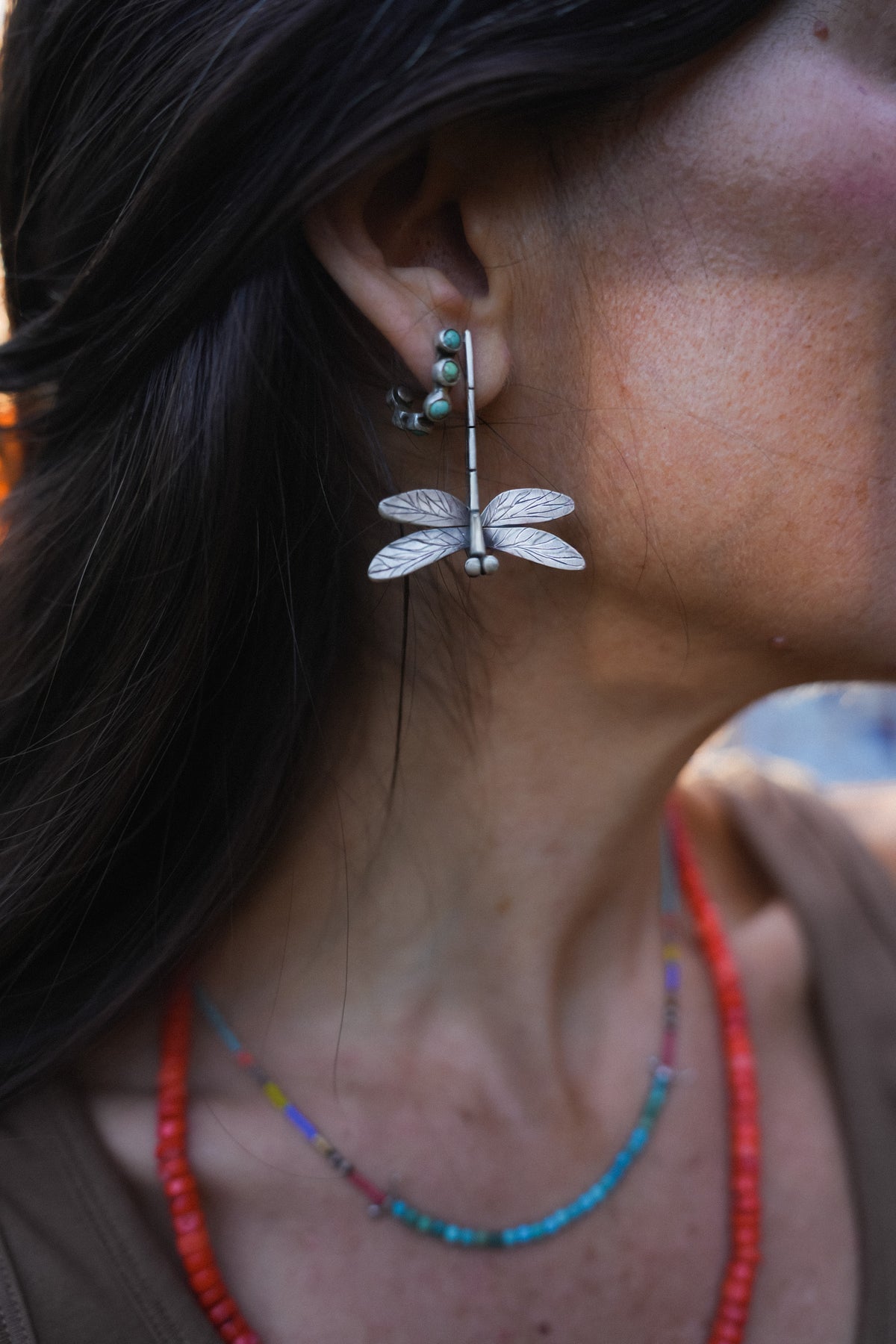 A close-up side profile of a woman wearing a large silver dragonfly dangle earring. She also wears silver hoop earrings with small turquoise beads and layered beaded necklaces.