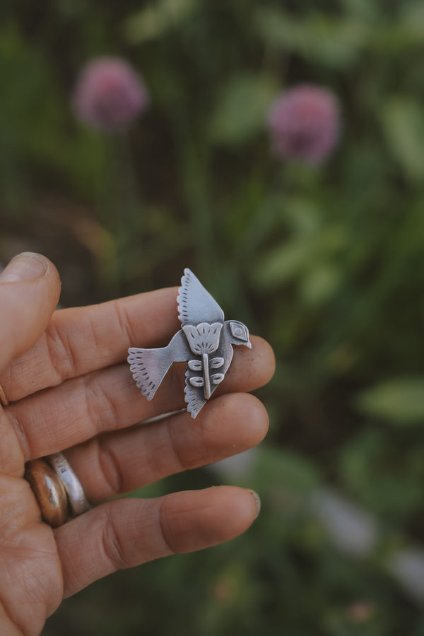 A woman's hand holding a small, etched, sterling silver Birkenstock shoe clip in the shape of a bird with outspread wings, against a blurred green background. The flower style is folk art.