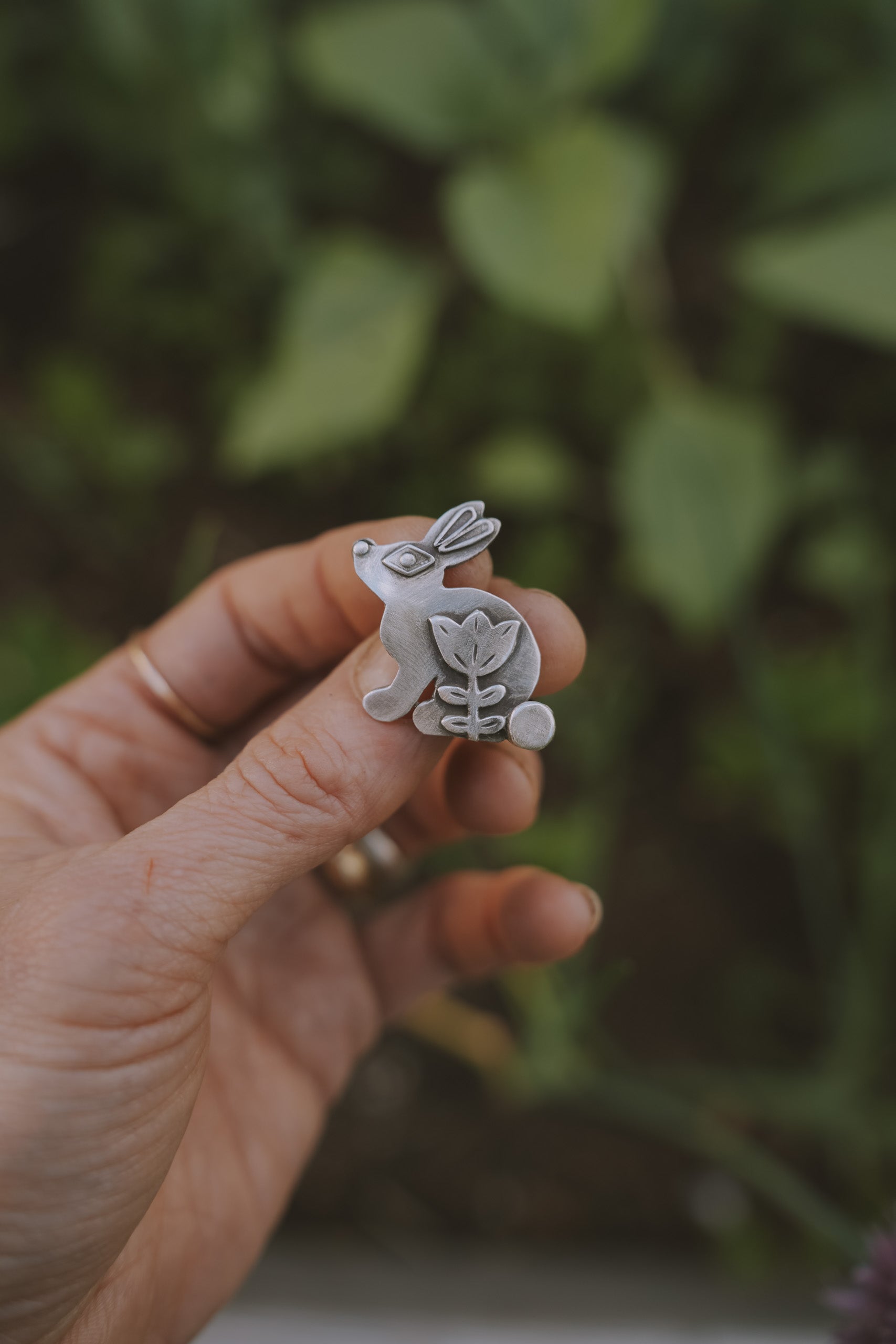 A woman's hand holding a small, etched, bunny-shaped sterling silver Birkenstock shoe clip against a blurred green background. The style is folk art.