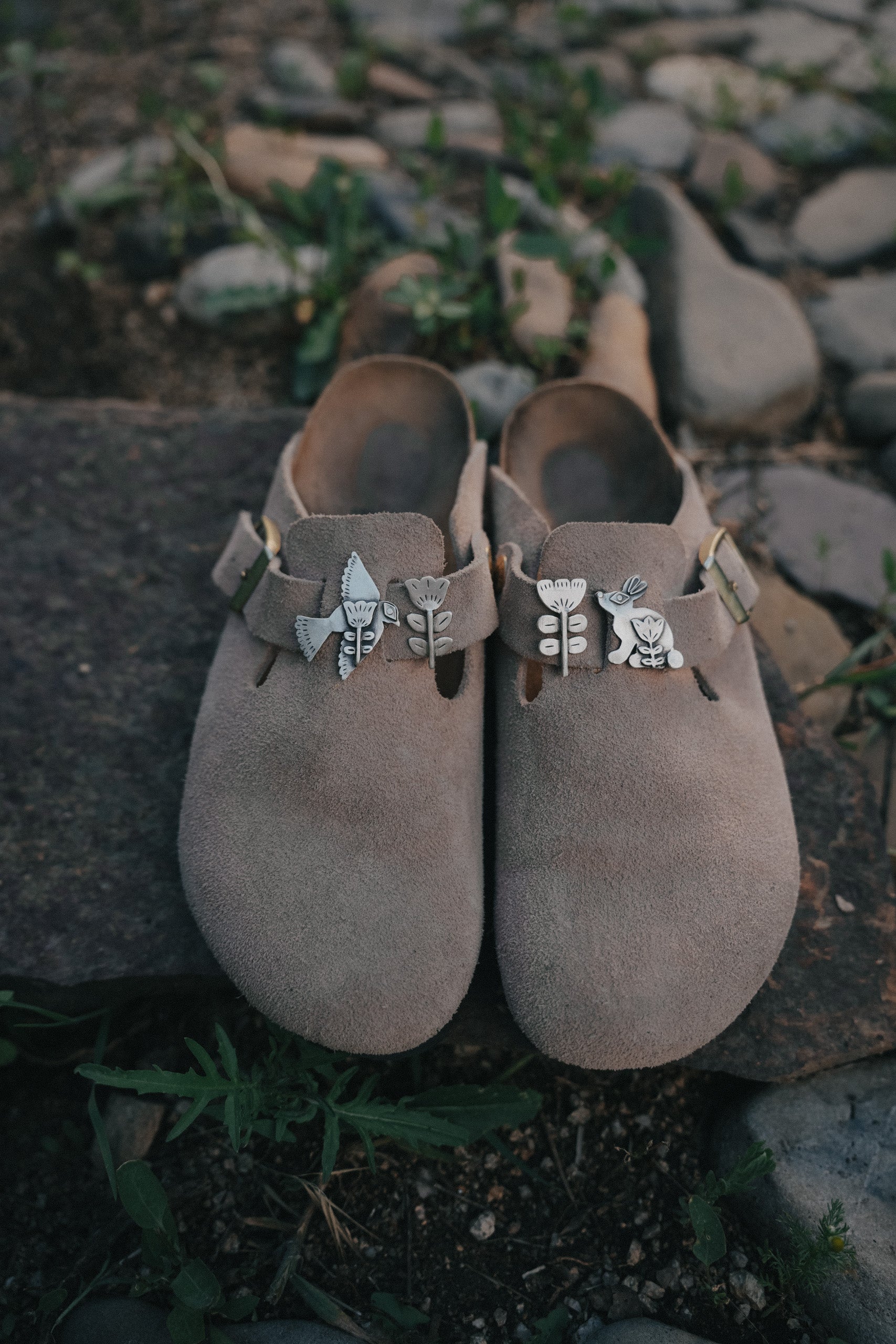 A pair of brown suede Birkenstock shoes with decorative sterling silver folk-styled shoe clips, set on a stone surface.