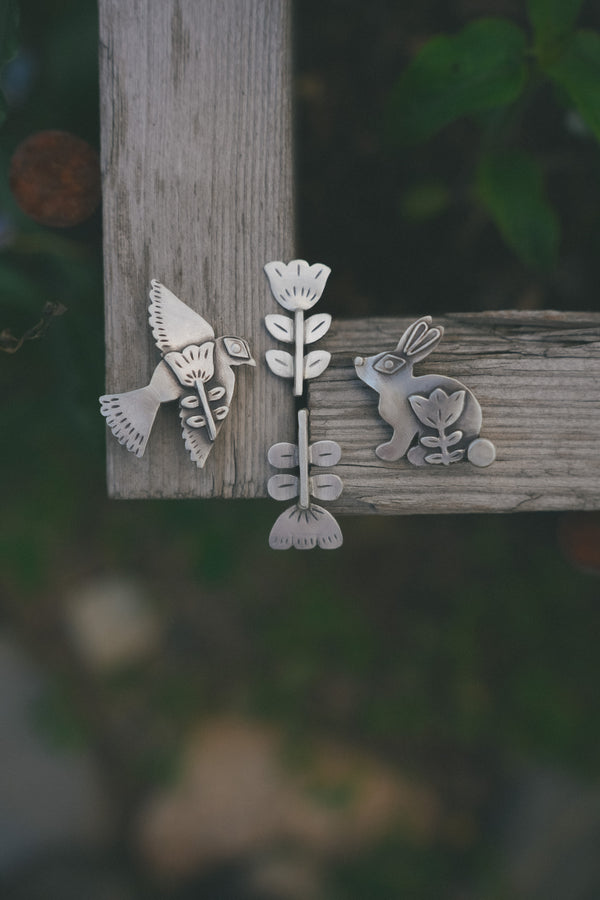 Three silver animal-shaped earrings on a wooden surface with a blurred natural background.