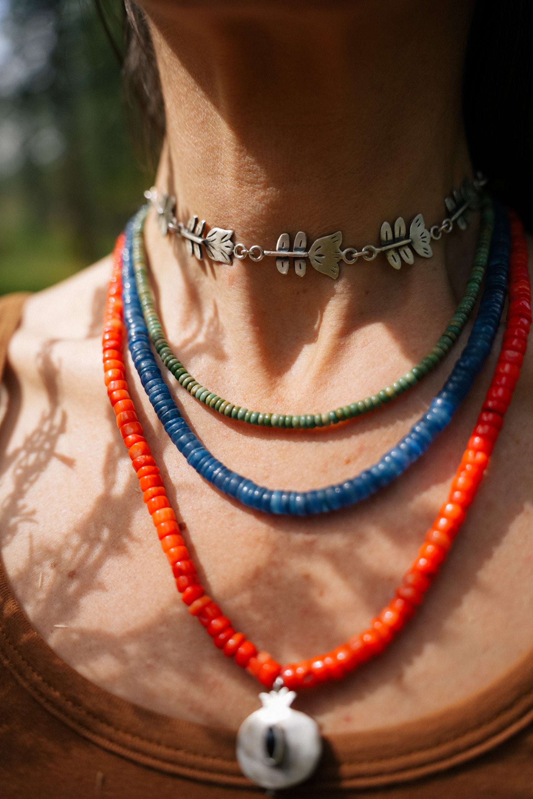 A close-up of a woman with dark hair wearing a sterling silver folk flower choker and three beaded necklaces. One is a slender strand of jade beads. The other two are this strands of larger lapis lazuli and coral. beads. 