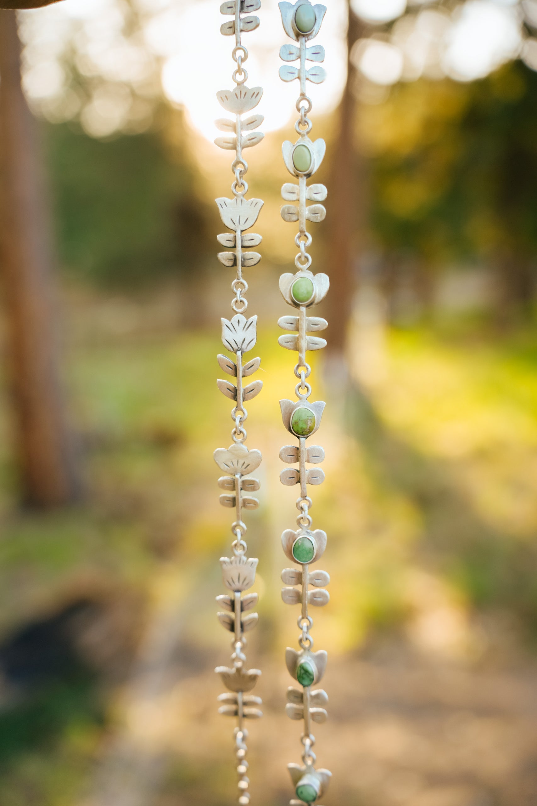Two sterling silver necklaces with folksy flower motifs suspended and hanging straight down with a sunny, blurred, natural background behind them. The flowers are simple and graphic in design - tulips and poppies, and a few other varieties. One of the chokers is all silver and the other has a variety of semi-precious gemstones adorning the flower heads.