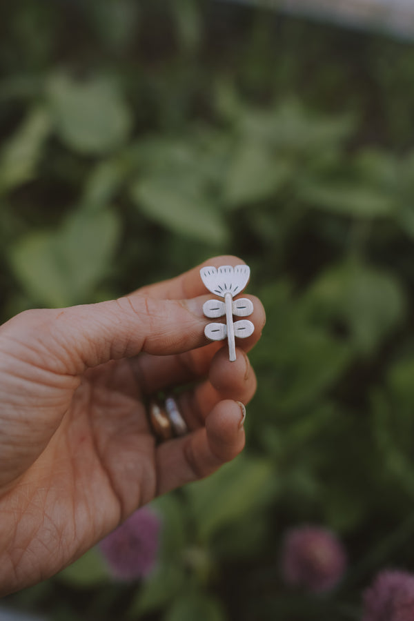 A woman's hand holding a small, etched, floral sterling silver Birkenstock shoe clip against a blurred green background. The flower style is folk art.