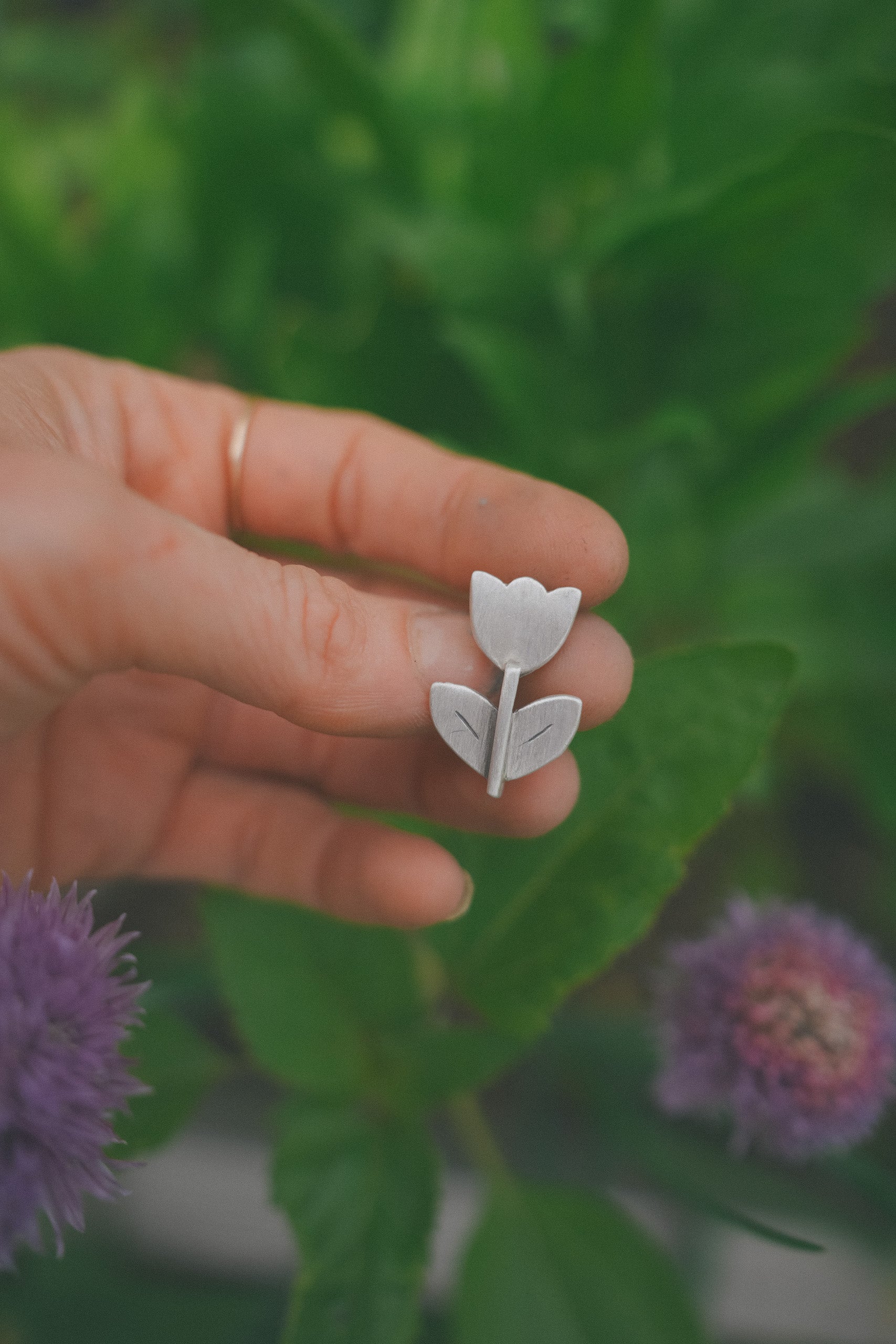 A woman's hand holding a small, etched, floral sterling silver Birkenstock shoe clip against a blurred green background. The flower style is folk art.