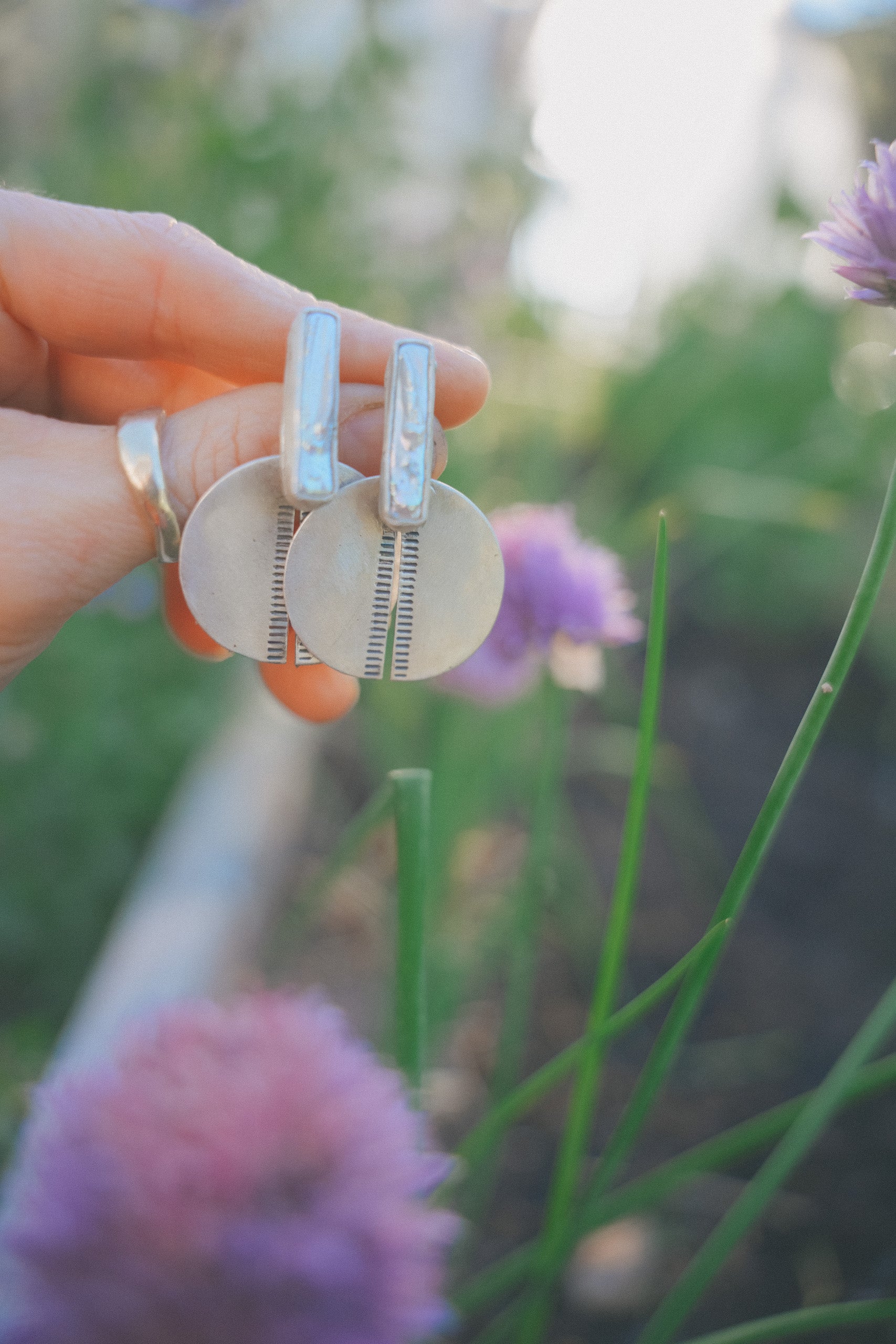 A close-up of a woman’s hand holding a pair of sterling silver and freshwater pearl earring, The freshwater pearls are narrow rectangular shapes set above split sterling silver discs with hand-stamping along the sides of the splits. The natural background is light green and lilac and is mostly out-of-focus.