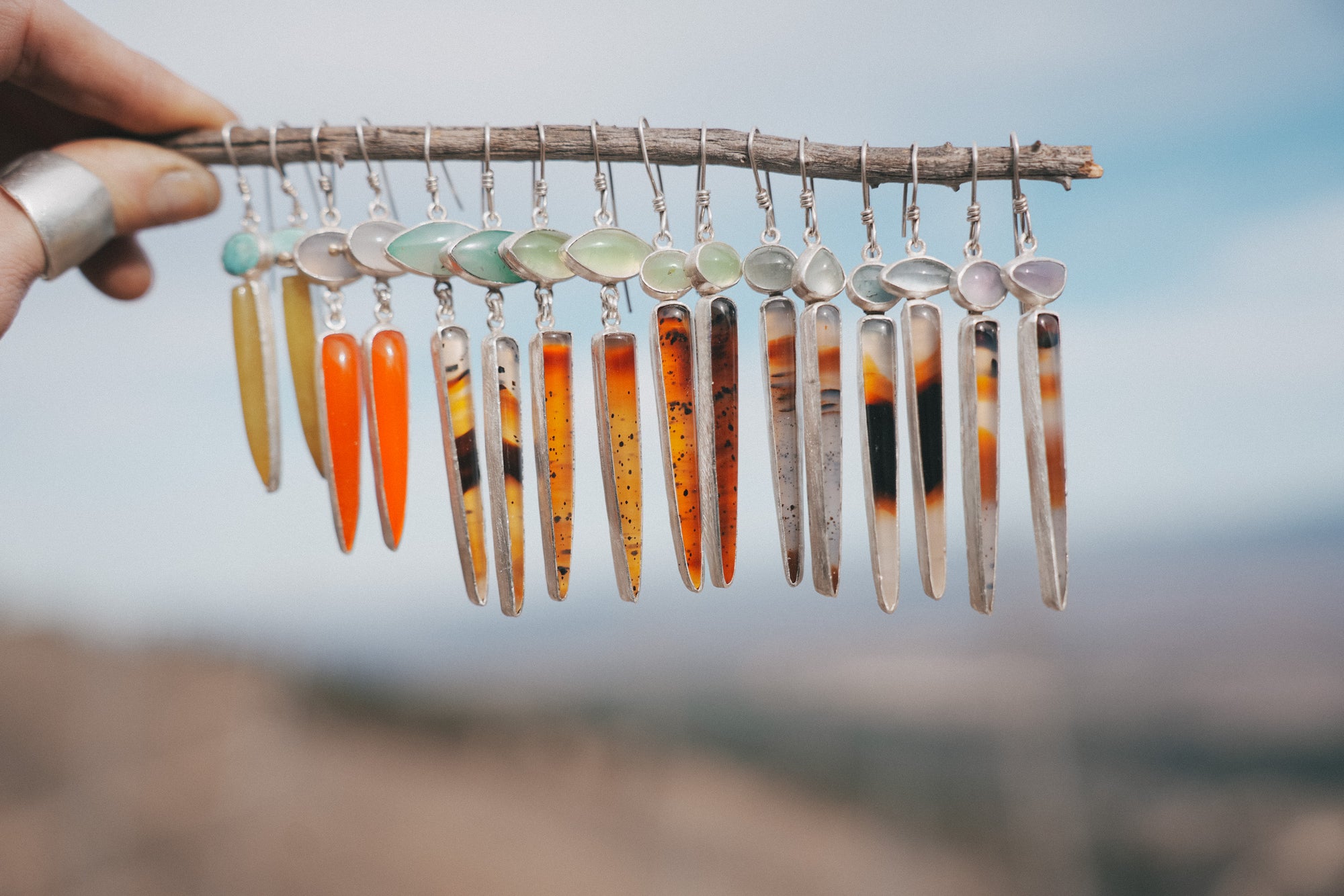 A woman's hand holding a branch with eight pairs of colorful sterling silver and spike-shaped gemstone earrings hanging from it against a blurred natural background.