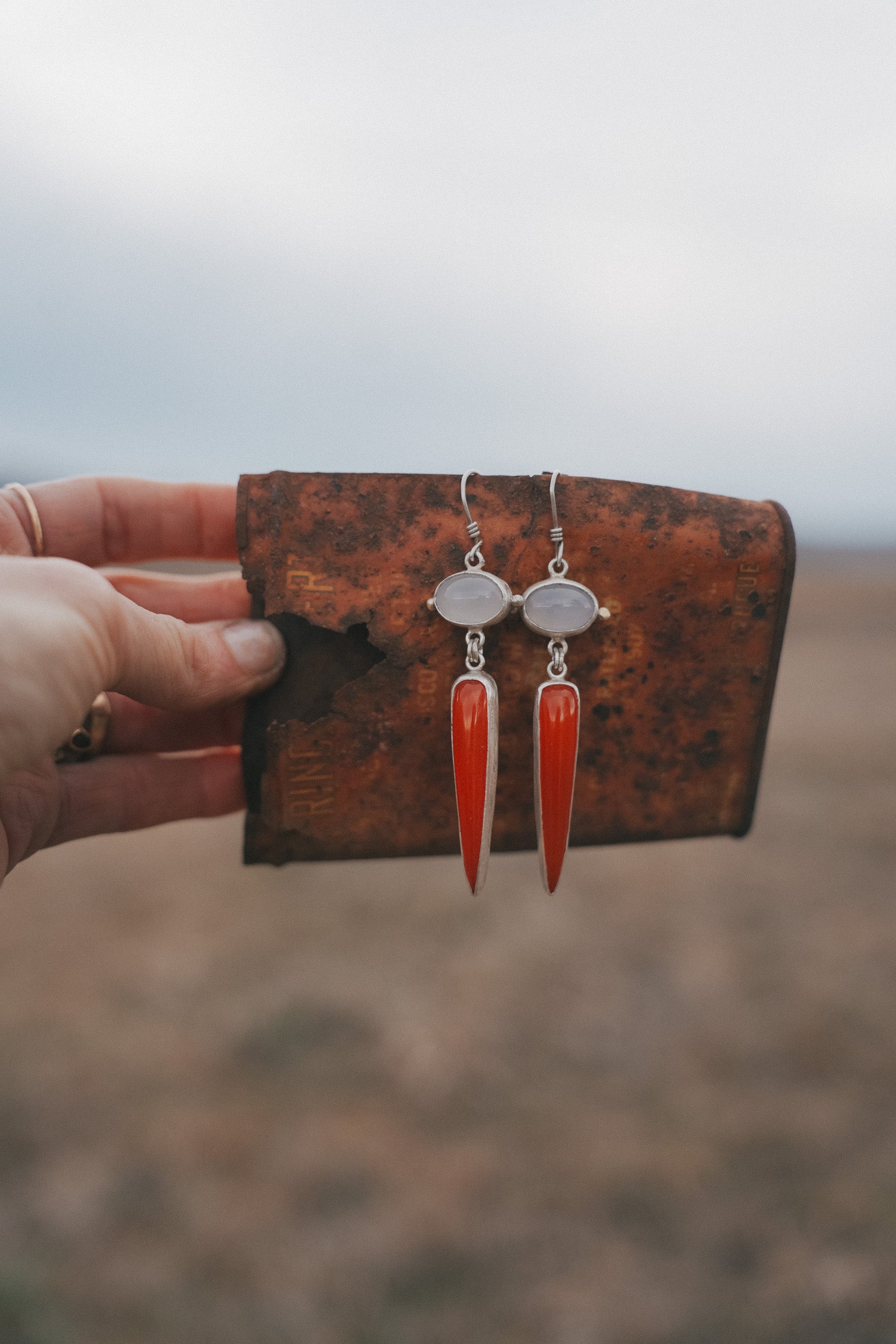 A woman's hand holding a pair of sterling silver, lilac-and-flame chalcedony drop earrings with 18K gold accents against a blurred natural background. The earrings are hanging from a flat, rusty can. The lilac chalcedony gemstones are oval, and the flame chalcedony spikes are a deep orange-red color.