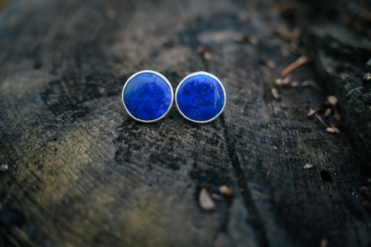 A pair of round stud earrings with vibrant blue lapis lazuli stones in silver bezel settings, resting on a dark textured wooden surface.