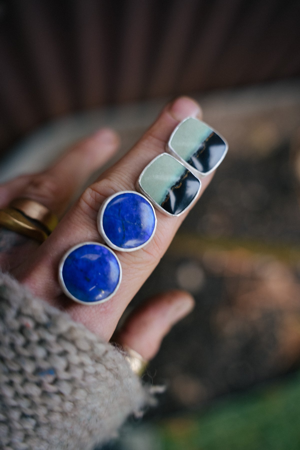 A pair of rectangular post earrings. They are sterling silver earrings set with chrysocolla gemstones, resting on a woman's hand. The gemstones are dark on the bottom and light to white on the top. A thin, organic ochre line runs horizontally across the stones, separating the dark from the light. She is also holding a pair of round lapis lazuli earrings.