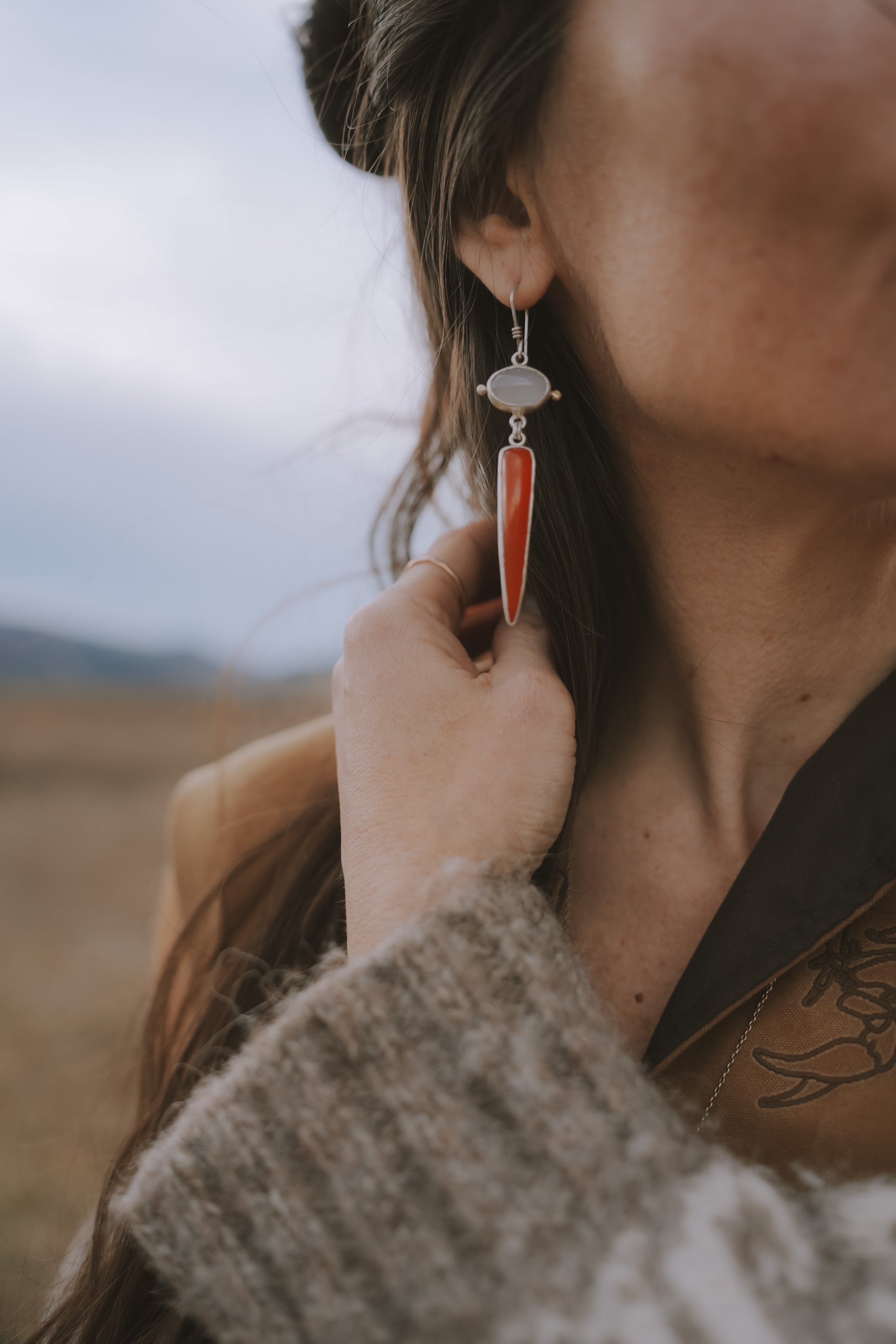 A woman wearing a pair of sterling silver, lilac and flame chalcedony drop earrings against a blurred natural background. She is pulling her long, dark hair back to show the earring. The lilac chalcedony gemstone is oval, and the flame chalcedony spike is a deep orange-red color.