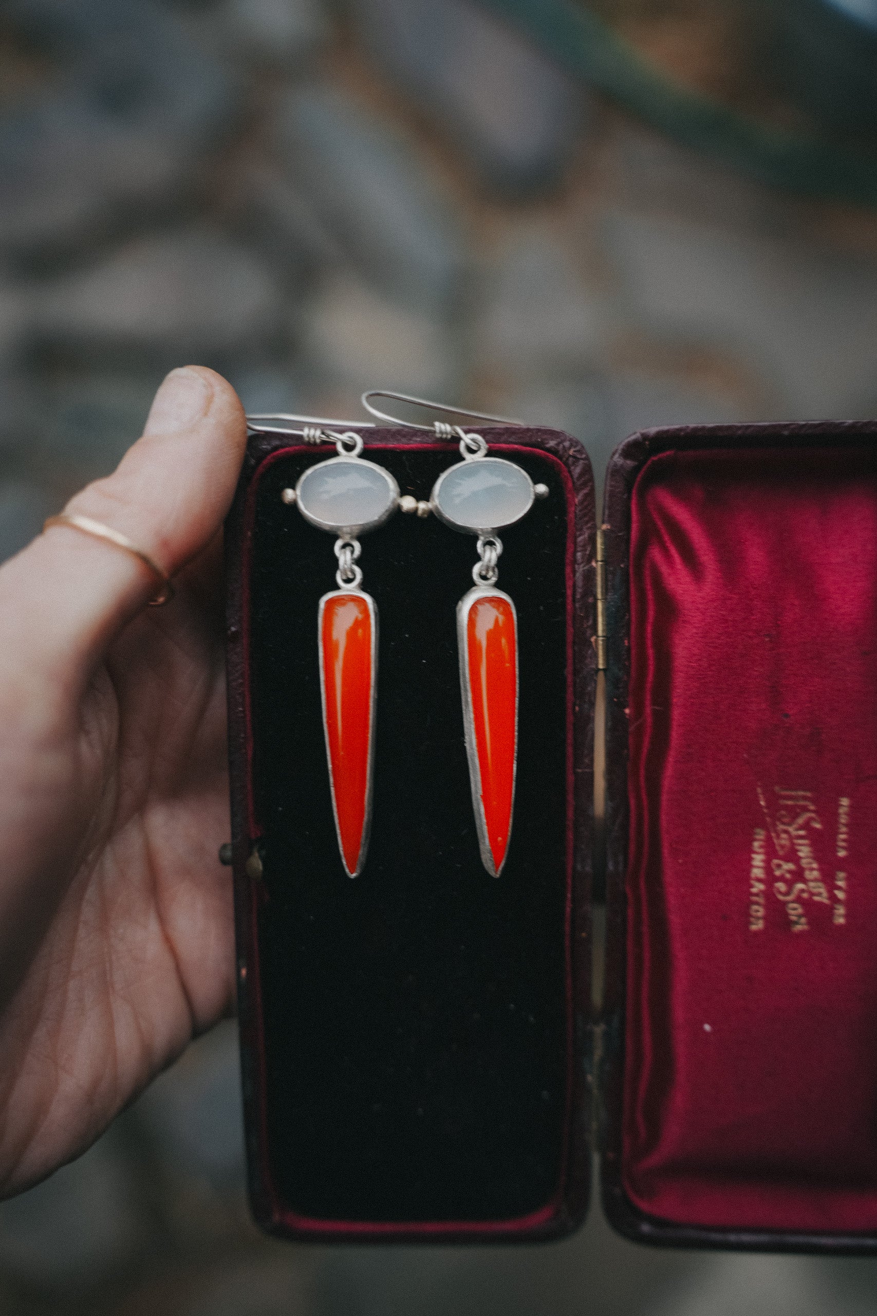 A woman's hand holding a pair of sterling silver, lilac and flame chalcedony drop earrings  set inside of an antique eyeglass case against a blurred natural background. The earrings are hanging from the top of the case. The lilac chalcedony gemstones are oval, and the flame chalcedony spikes are a deep orange-red color.