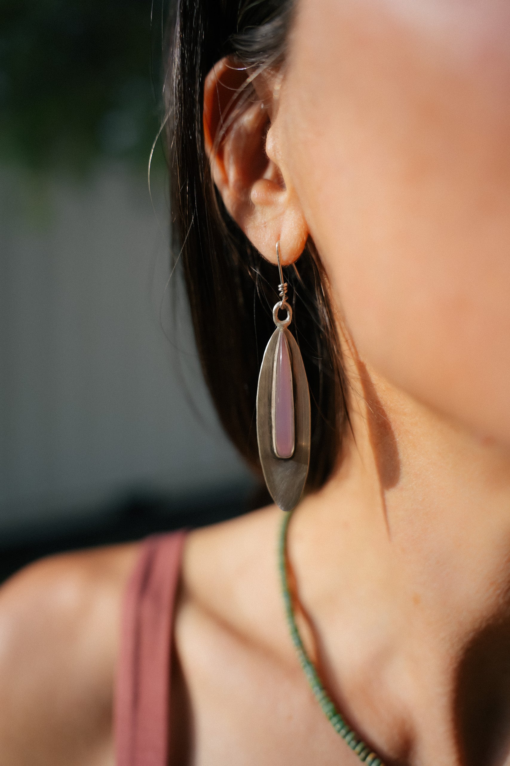 Close-up of a woman with dark hair wearing a sterling silver marquise-shaped, dangler earring and an elongated llilc-colored chalcedony gemstone set in the center. The background is blurred.