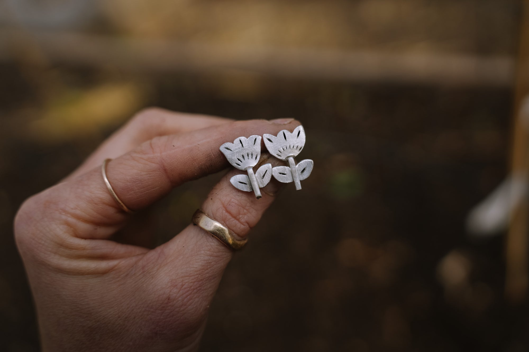A woman's hand holding a pair of stamped and carved sterling silver tulip earrings against a blurred natural background. The tulips are simple and graphic.