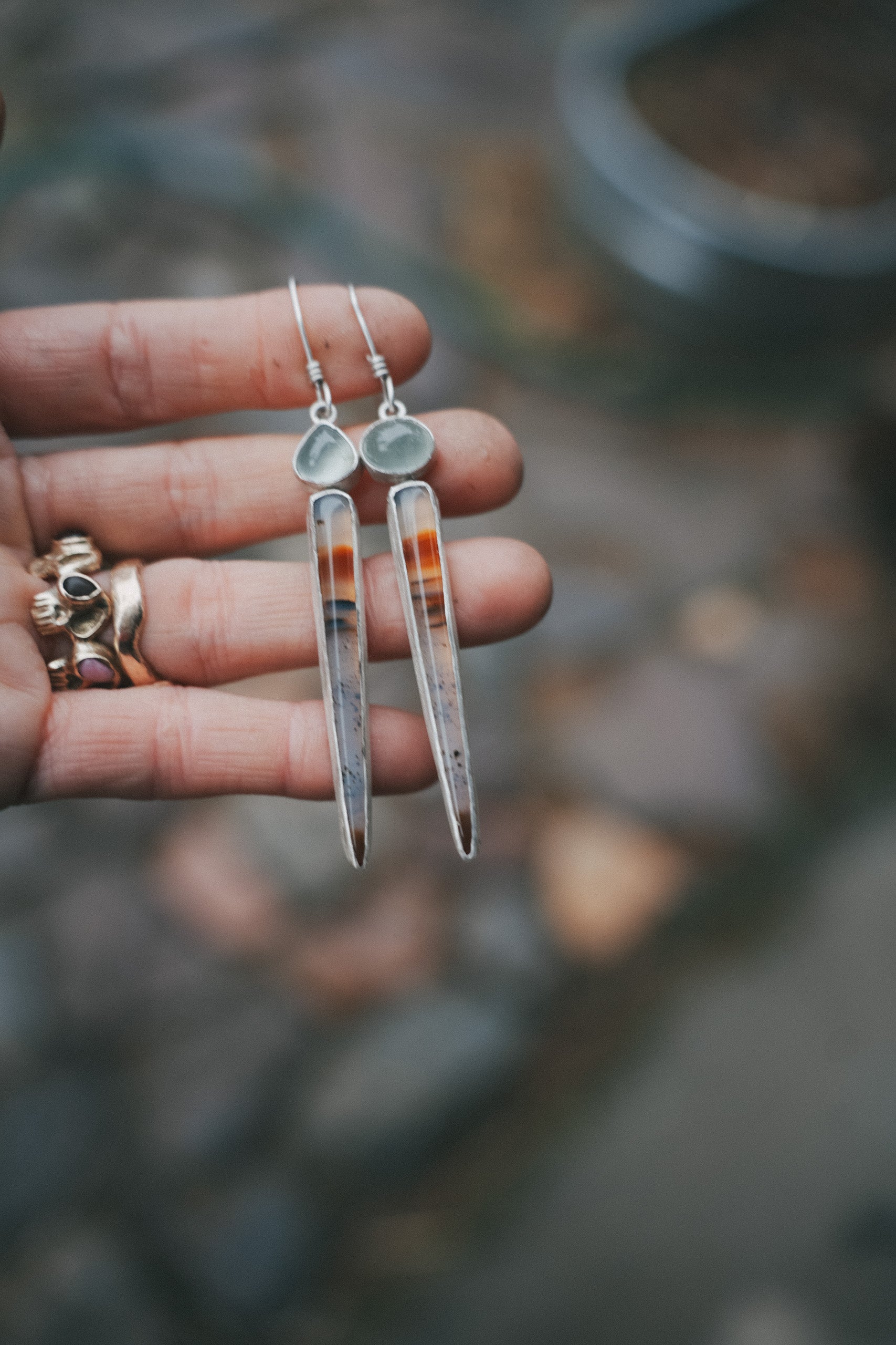 A woman's hand holding a pair of sterling silver, Montana agate, and aquamarine drop earrings against a blurred natural background. The oval aquamarine gemstones are pale blue-green, and the Montana agate spikes have bands of red-orange and dark brown with black specs throughout.