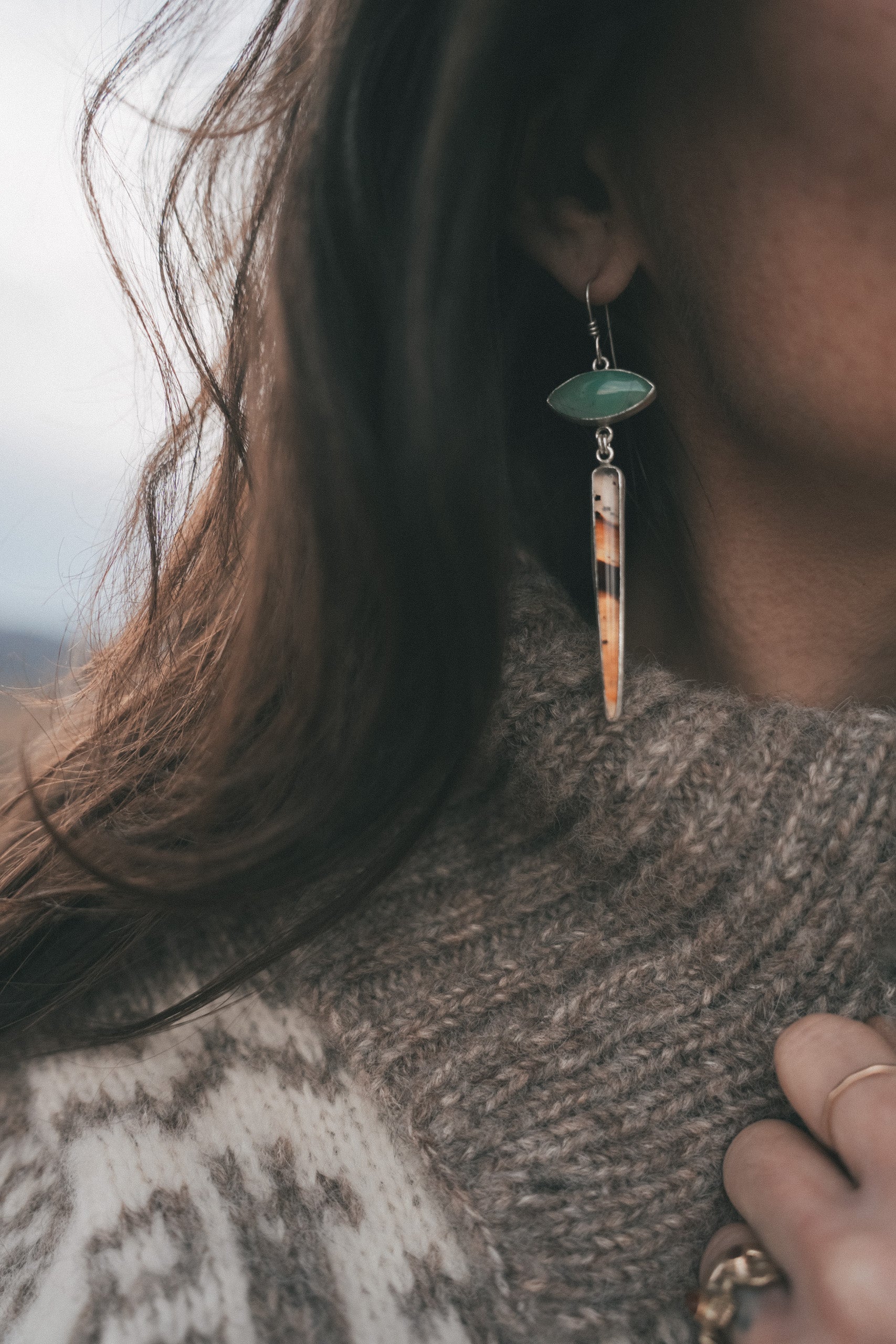 A woman wearing a sterling silver, Australian chrysoprase and Montana agate drop earrings against a blurred natural background. She has long, dark hair back is wearing a thick sweater. The Australian chrysoprase gemstone is pale green and eye-shaped and the Montana agate spike has bands of orangey caramel, dark brown-black, and cream colors with black flecks.