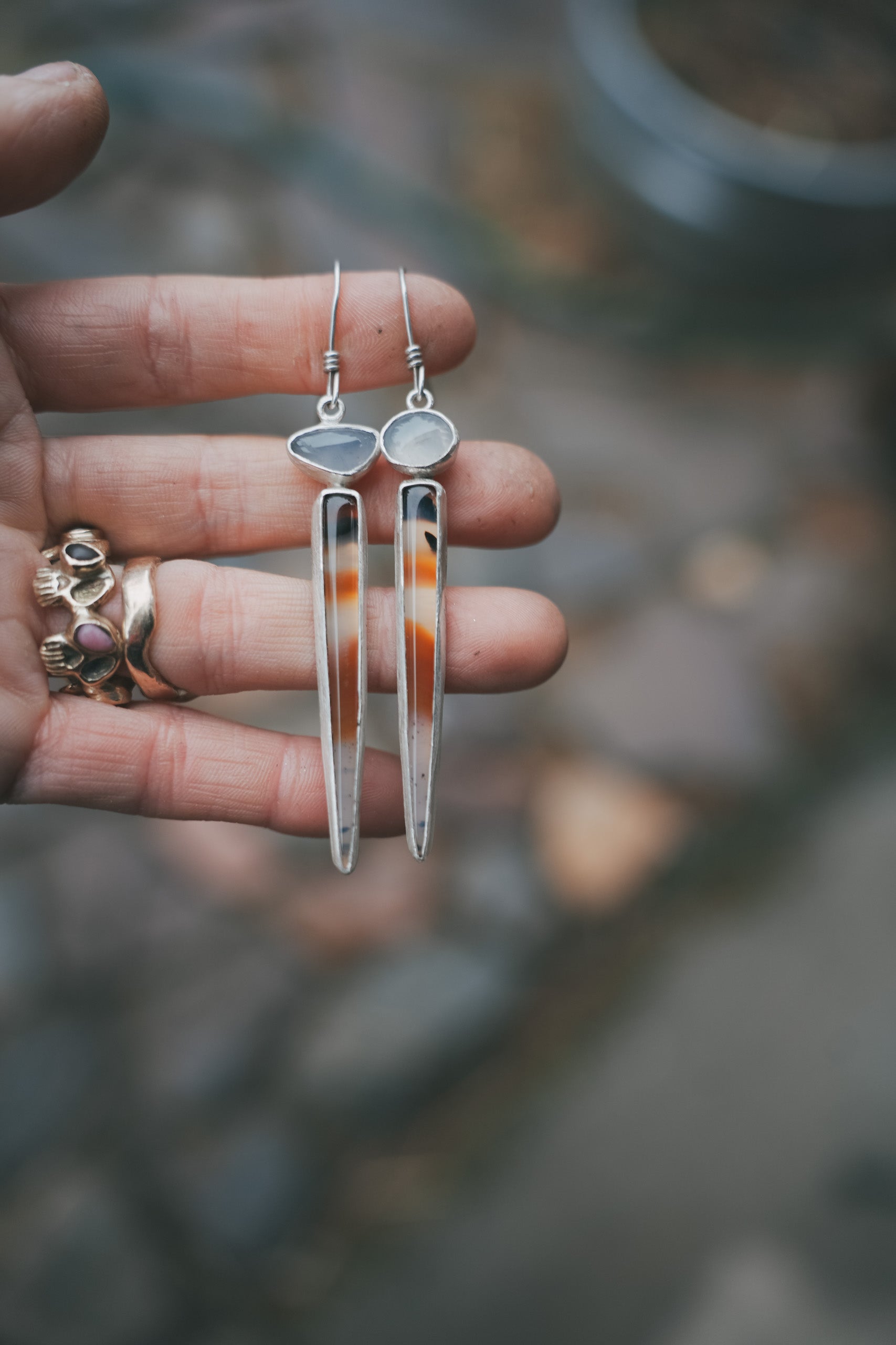A woman's hand holding a pair of sterling silver, Montana agate, and lilac chalcedony drop earrings against a blurred natural background. The oval chalcedony gemstones are pale lilac, and the Montana agate spikes are an orangey caramel and dark brown-black color with black flecks in a semi-clear milky base.