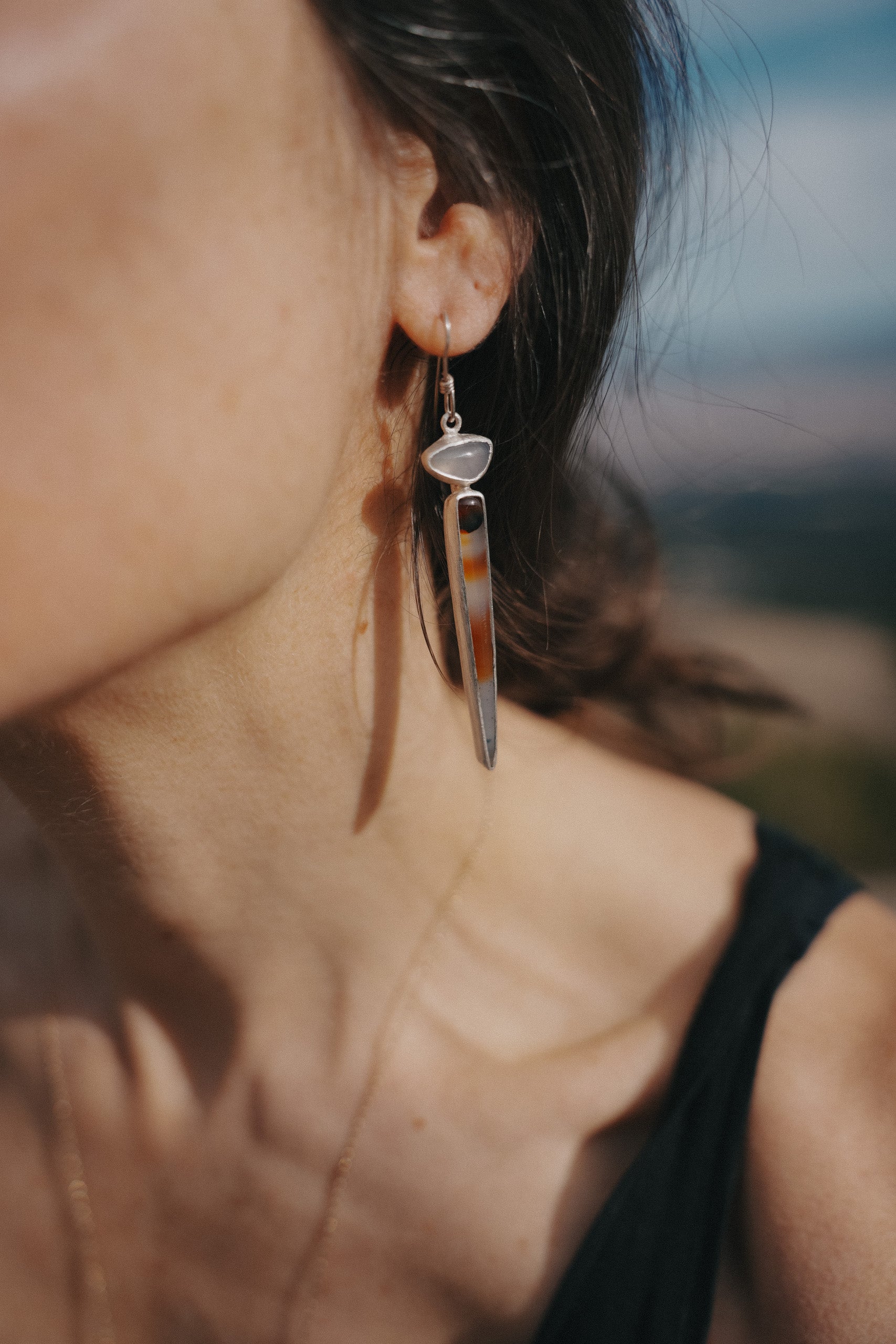 A woman wearing a sterling silver, lilac chalcedony and Montana agate drop earrings against a blurred natural background. Her long, dark hair back is pulled to show the earring. The lilac chalcedony gemstone is an irregular oval, and the fMontana agate spike is an orangey caramel and dark brown-black color with black flecks in a semi-clear milky base.