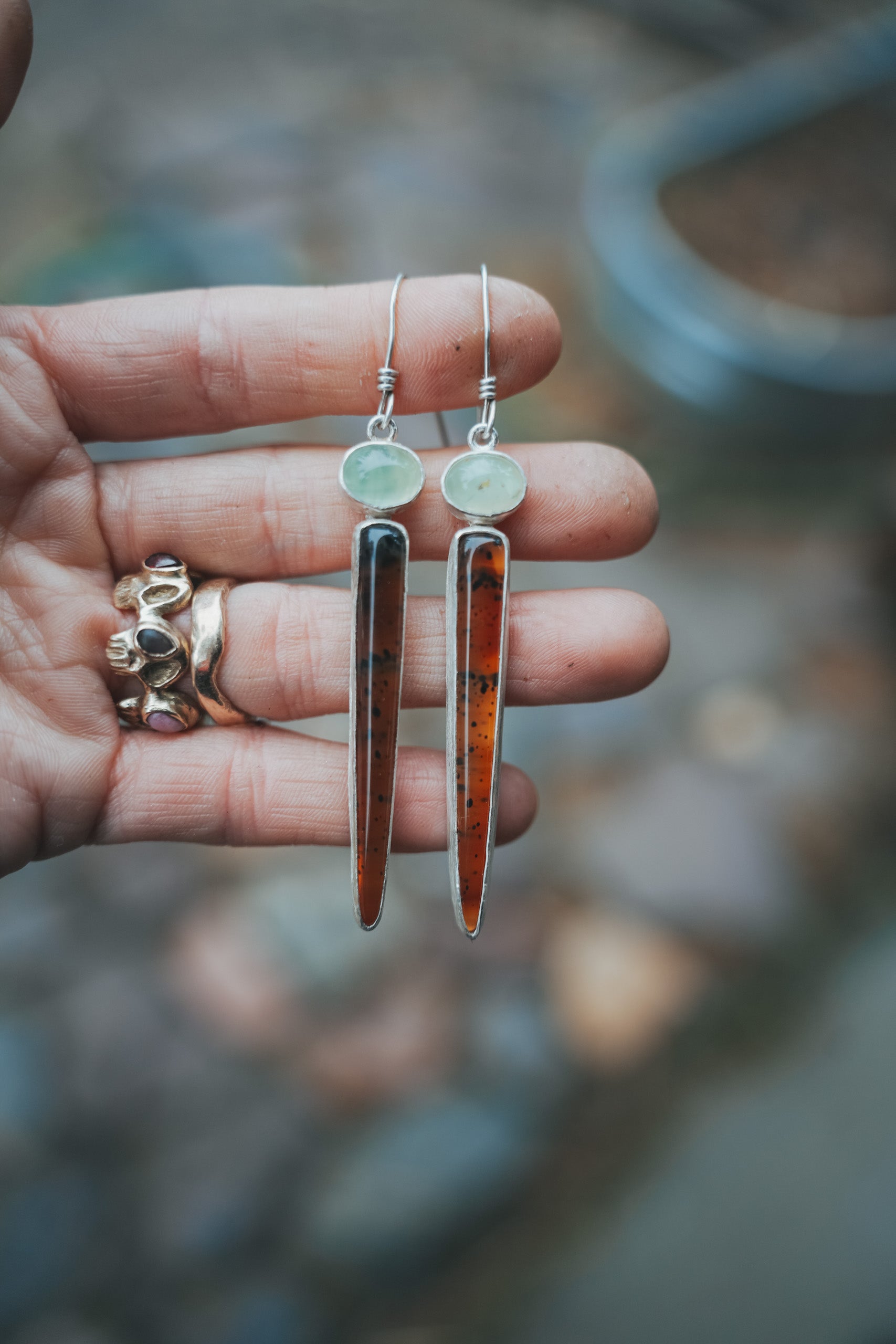 A woman's hand holding a pair of sterling silver, prehnite, and Montana agate drop earrings against a blurred natural background. The oval prehnite gemstones are pale green, and the Montana agate spikes are an orangey caramel color with black flecks throughout.