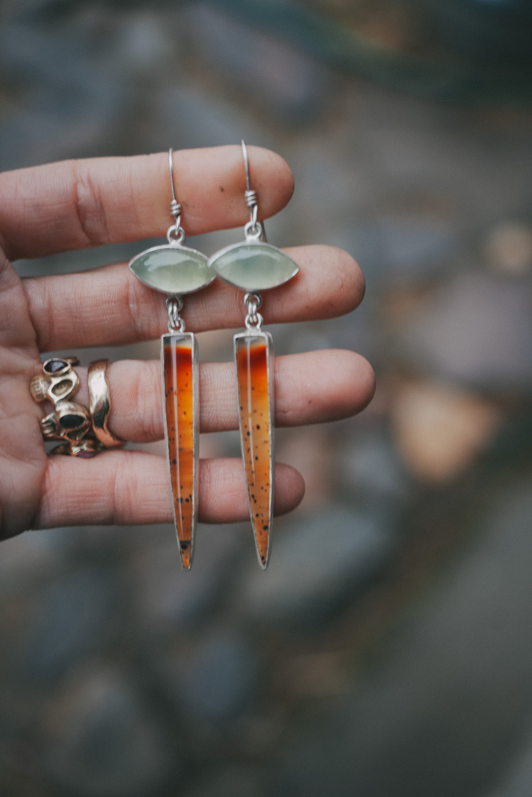A woman's hand holding a pair of sterling silver and gemstone earrings against a blurred natural background.
The pale green prehnite gemstones at the top are eye-shaped, and the speckled orange Montana agate gemstones at the bottom are spike-shaped.