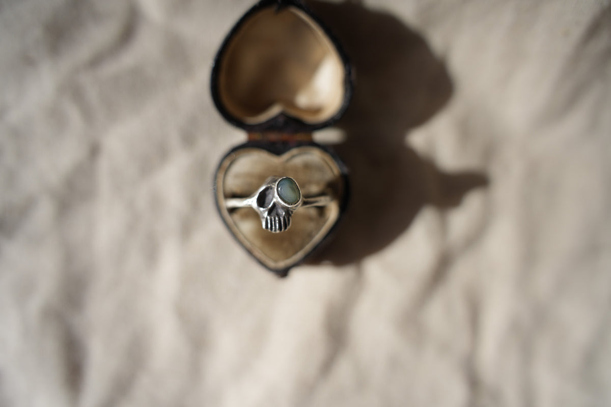 A silver skull ring detailed with an opal, displayed inside an open heart-shaped ring box on a textured beige fabric background.