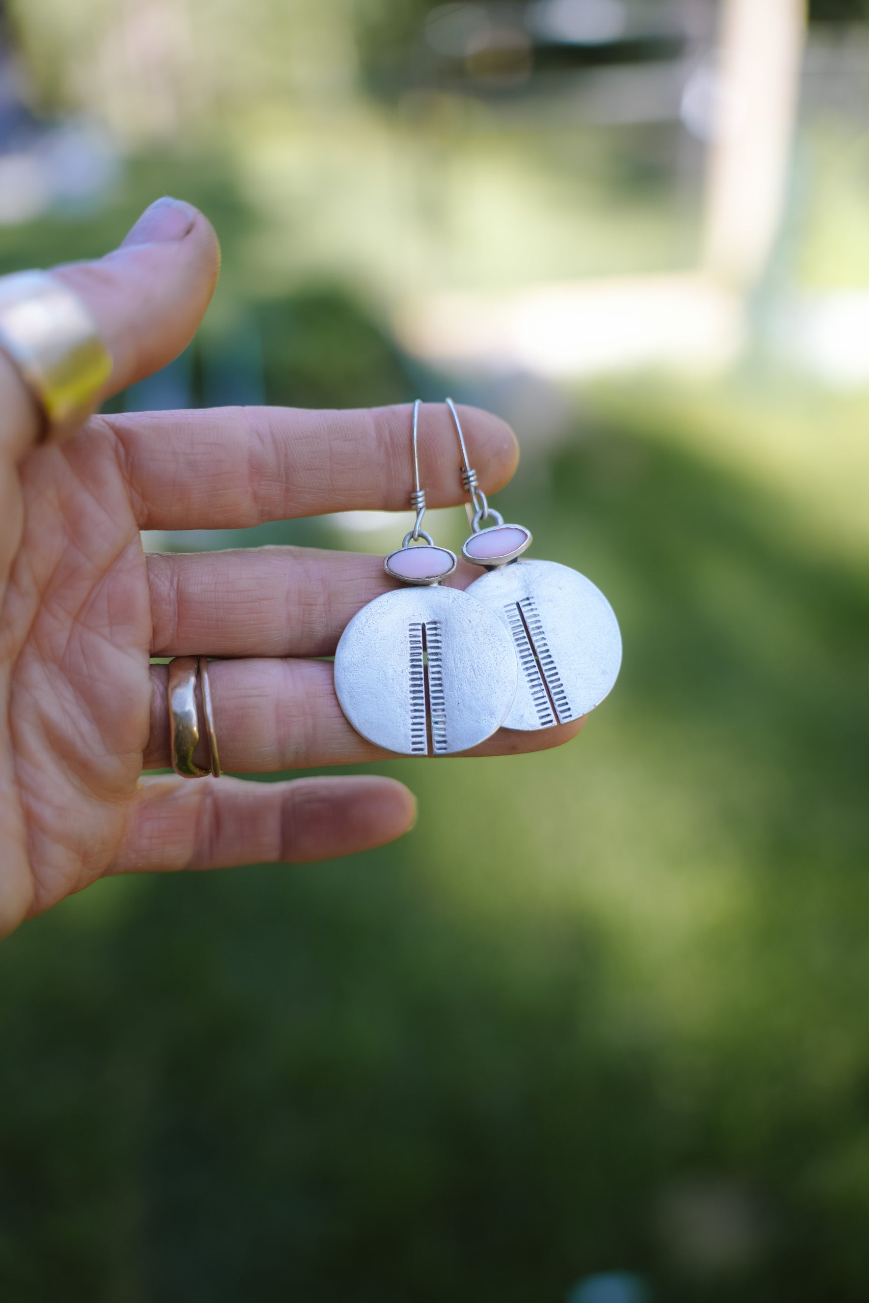 A woman's hand holding a pair of handmade silver earrings with oval pink opal gemstones set at the top. The sterling silver discs are split in the middle with etched vertical lines on each side of the split. Set against a blurred green outdoor background.