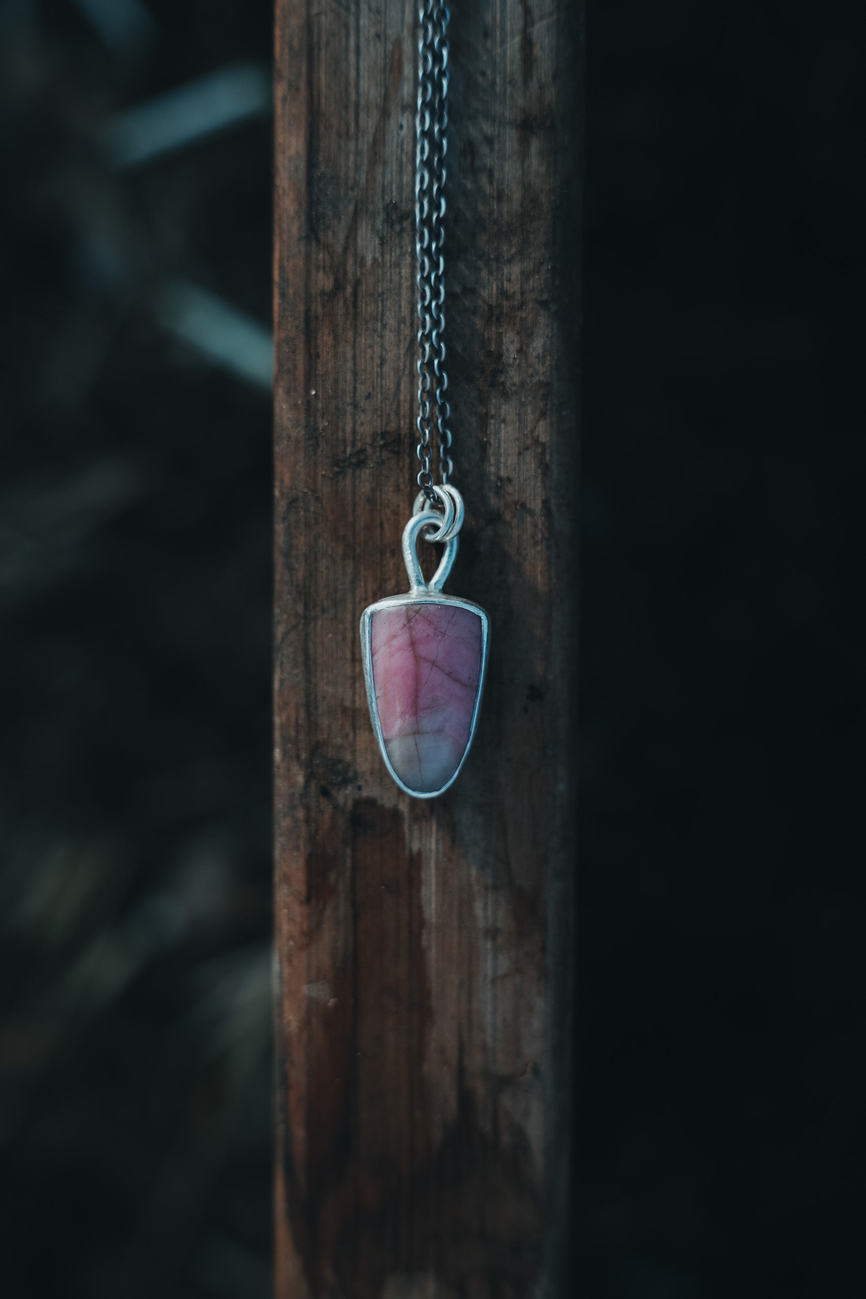 A sterling silver and pink opal shield-shaped pendant on a sterling silver chain hanging in from of a weathered wood post.