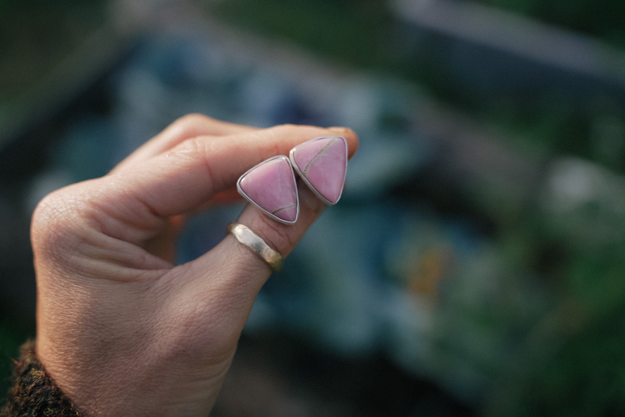 Hand holding two sterling silver and pink opal triangular earrings with a blurred background