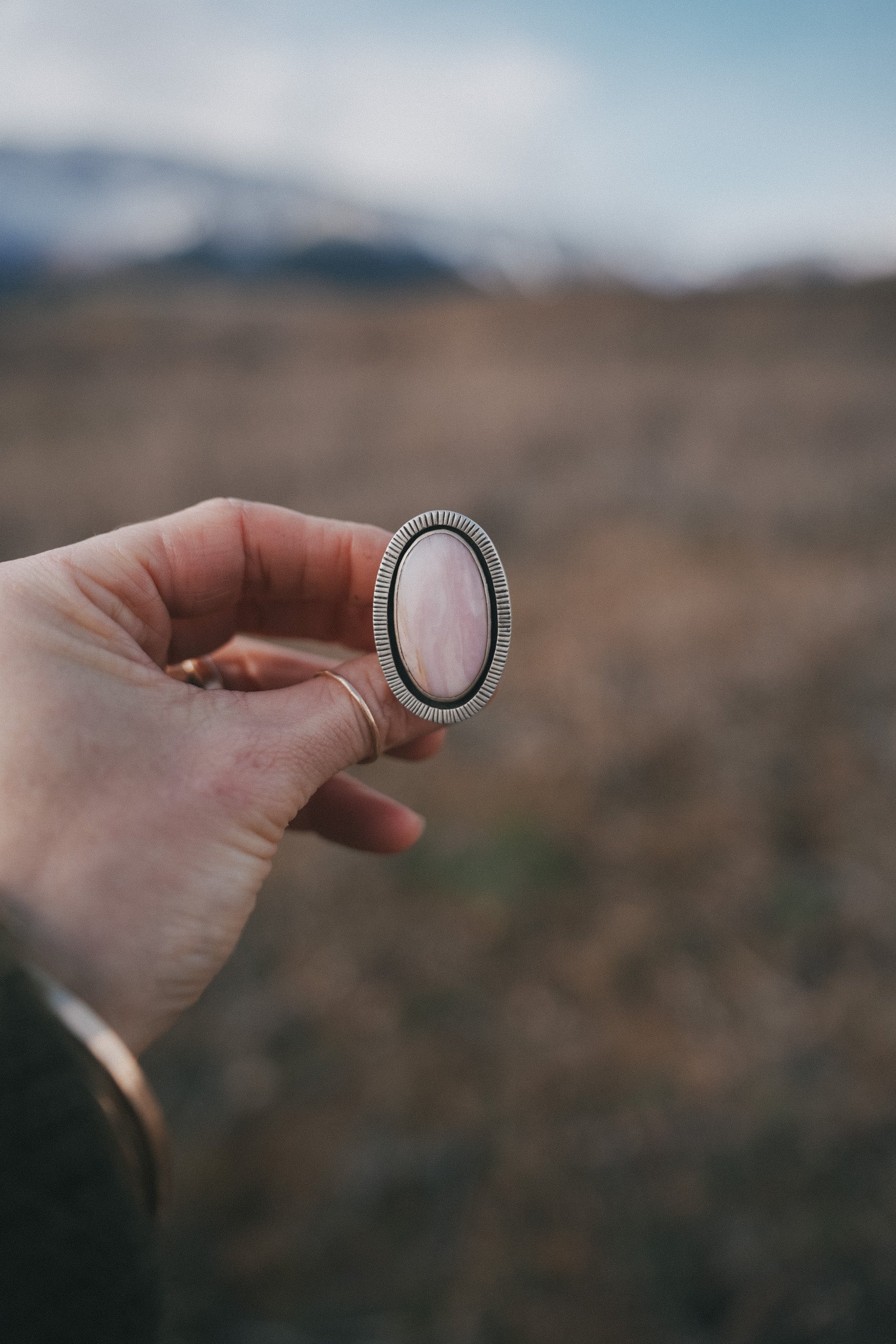 A woman's hand holding a sterling silver ring with a pink opal against a blurred natural background. The gemstone is oval-shaped, and there is stamping on the silver surrounding it.