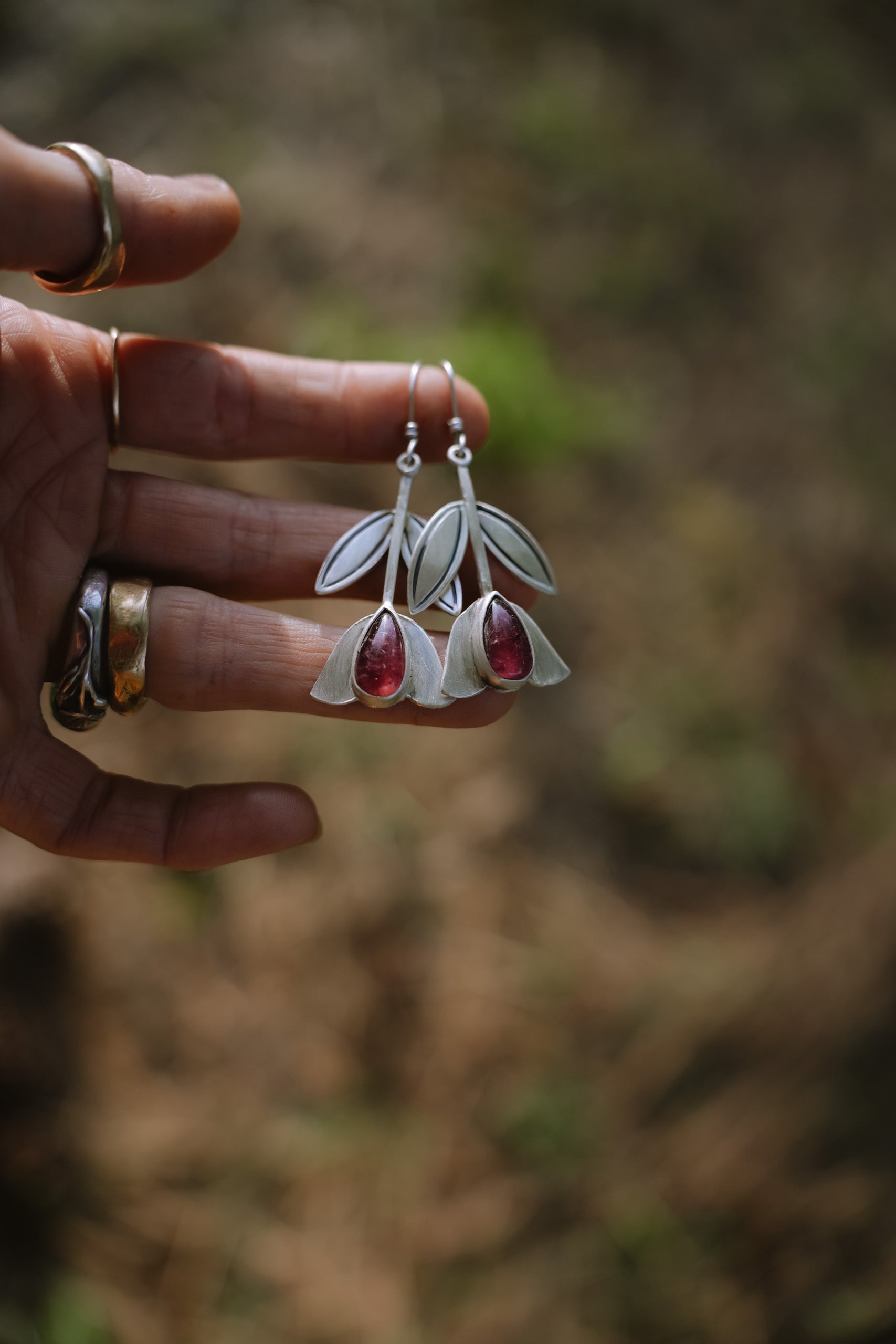 A woman's hand holding a pair of sterling silver dangler earrings with pink tourmaline gemstones. The gemstones are tear-drop-shaped. The earrings are shaped like tulips. The natural background is blurred. 