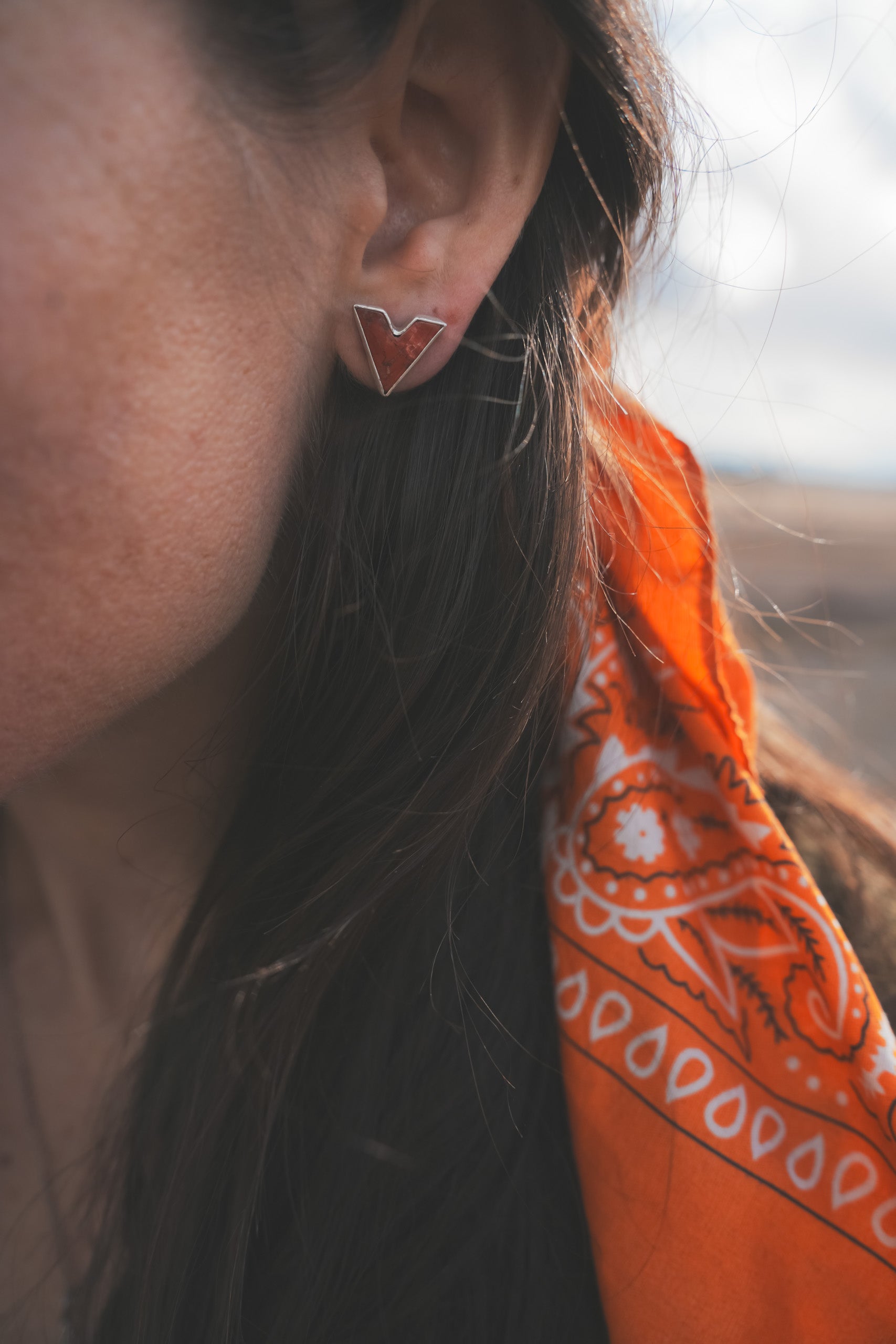 Close-up of a woman wearing a sterling silver and red poppy jasper V-shaped post earring. She is outside wearing an orange bandanna, and the background is blurred.