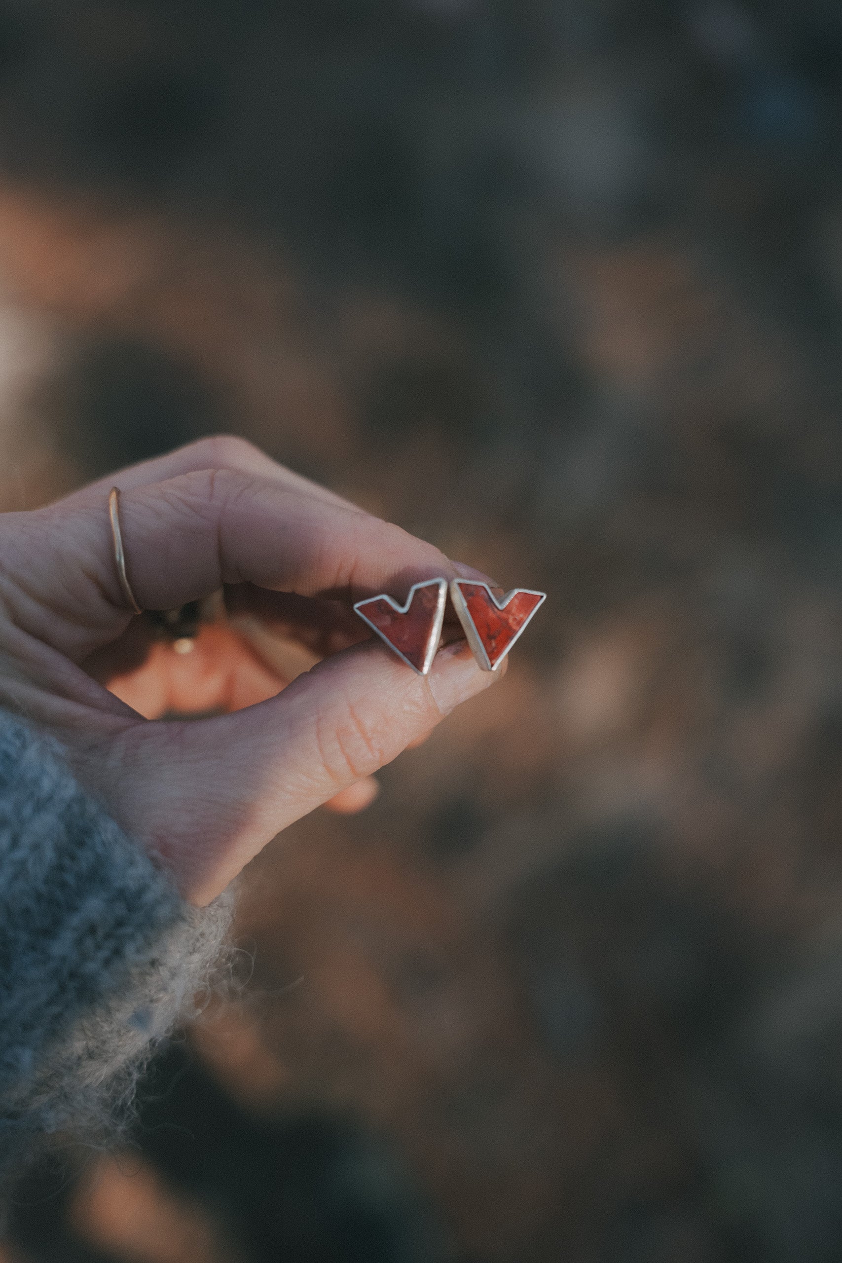 A woman's hand holding a pair of V-shaped sterling silver and poppy jasper post earrings against a blurred natural background.