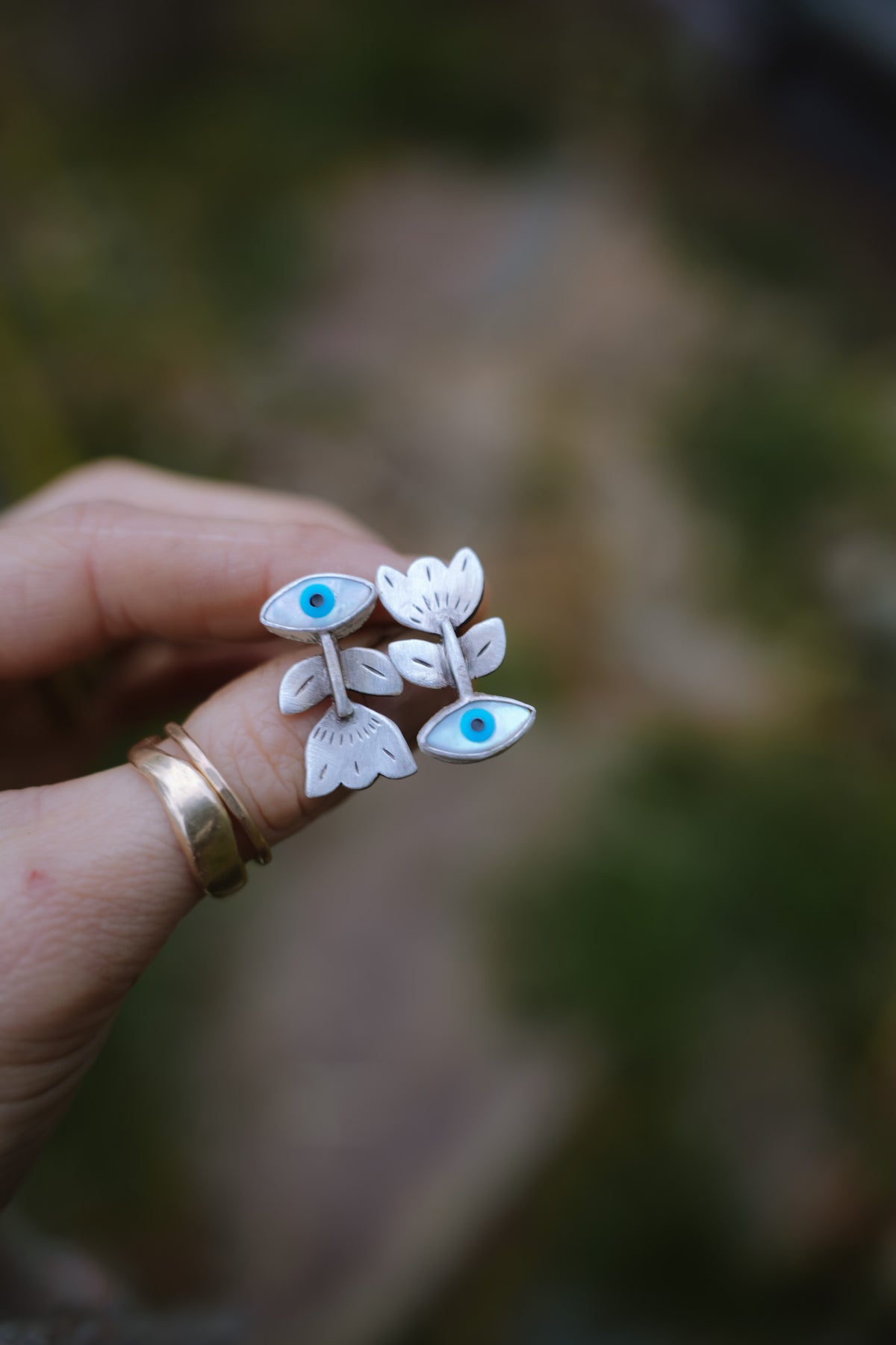 A woman is holding a pair of sterling silver post earrings with hand-stamped flowers and turquoise blue evil eye accents, set against a blurred outdoor background.