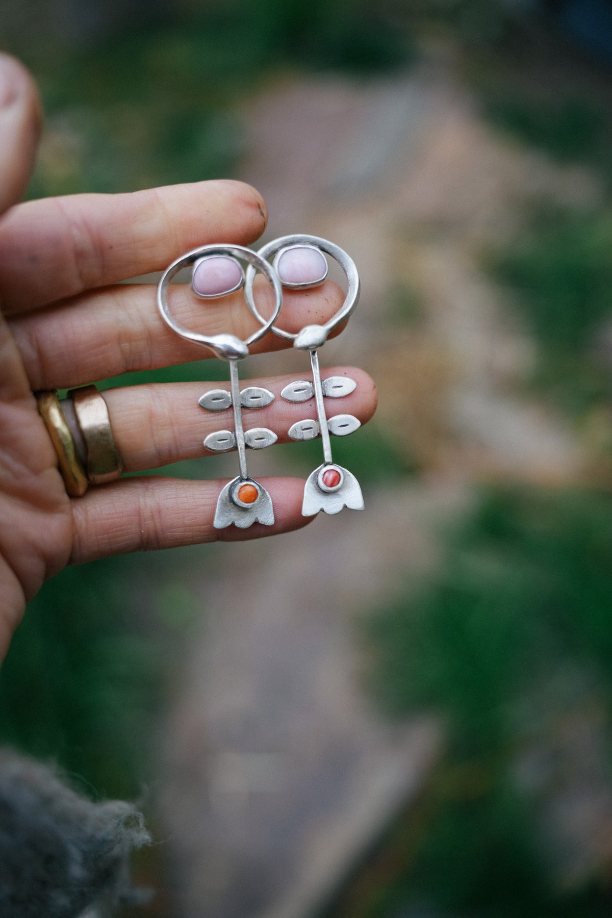 A woman's hand holding a pair of handmade silver earrings with light pink stones and small orange accent stones, against a blurred outdoor background. The earring design includes a snake and a folk-inspired flower.
