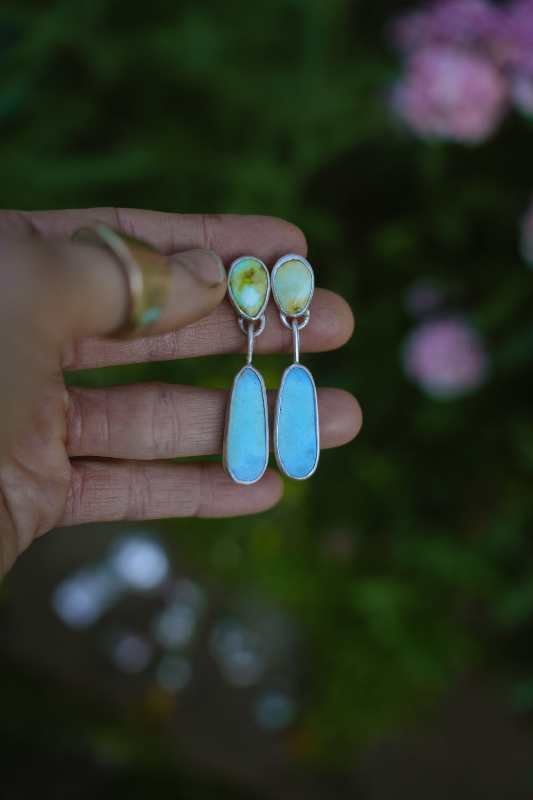 A woman's hand holding a pair of sterling silver and turquoise dangler earrings against a blurred green garden background with pink flowers. The earrings have teardrop-shaped light blue and green tops and elongated light blue turquoise ovals set in silver at the bottom.