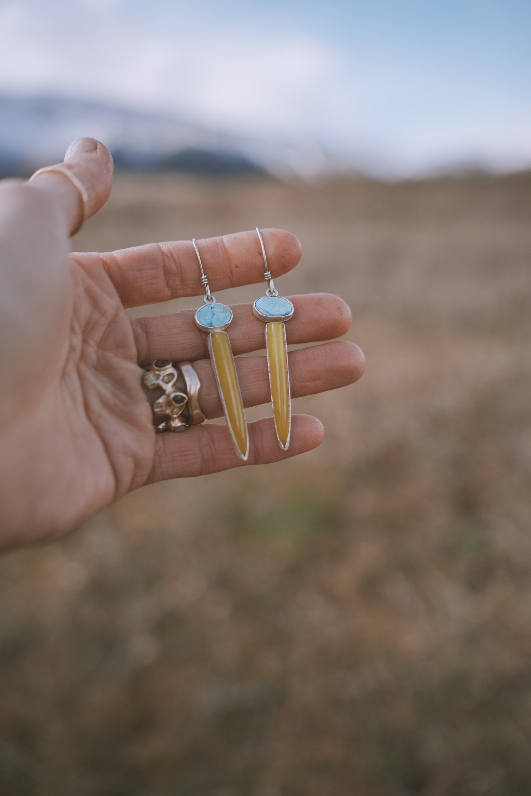 A woman's hand holding two sterling silver, turquoise, and vesuvianite drop earrings against a blurred natural background. The oval turquoise gemstones are pale blue, and the vesuvianite spikes are caramel-colored.