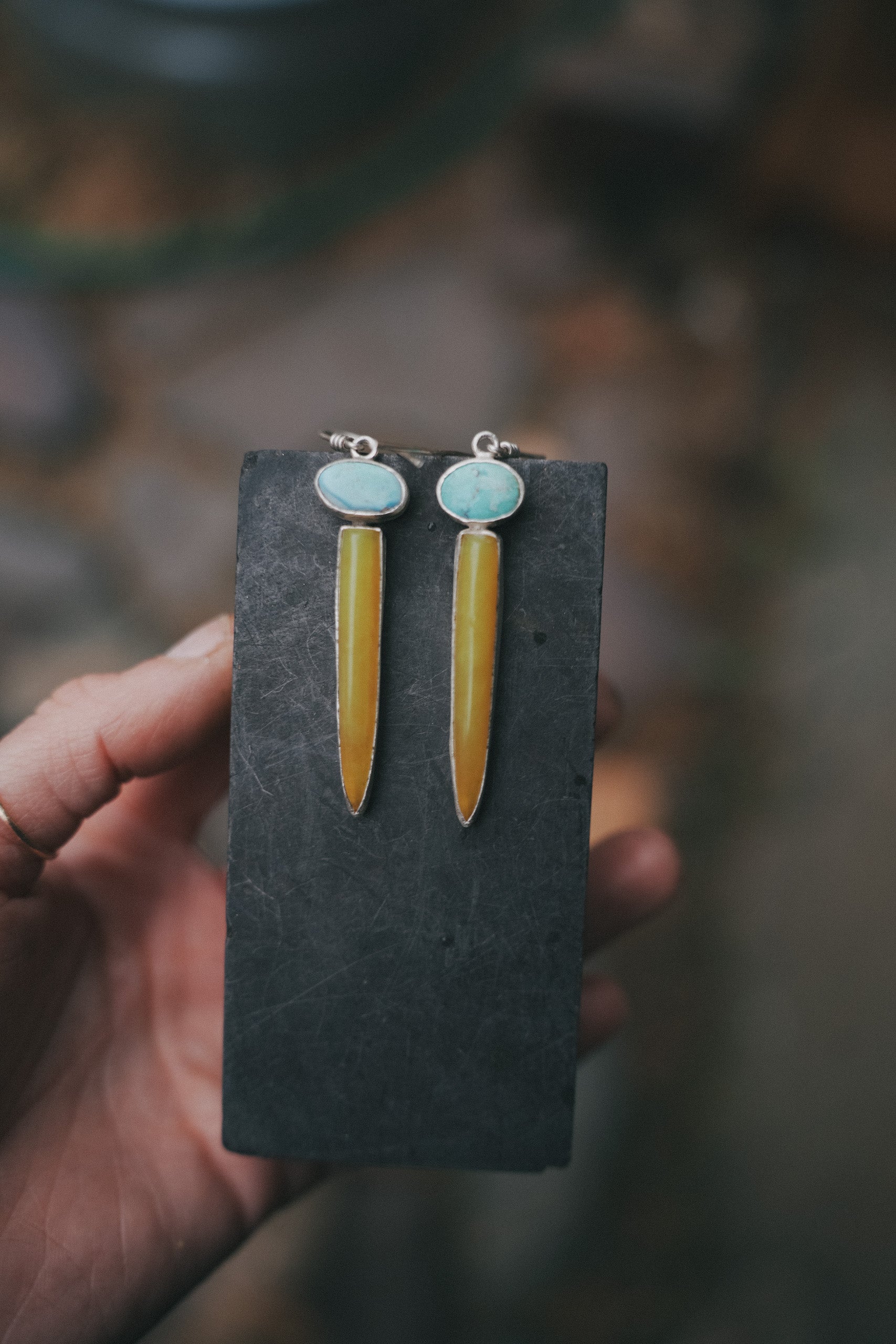A woman's hand holding a pair of sterling silver, turquoise, and vesuvianite drop earrings hanging from a dark piece of stone against a blurred natural background. The oval turquoise gemstones are pale blue, and the vesuvianite spikes are caramel-colored.